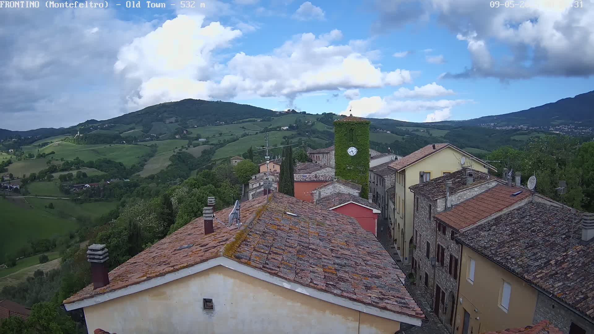 A partly cloudy day reveals a view of a village nestled in a verdant valley, with terracotta rooftops and a clock tower prominent among the buildings.