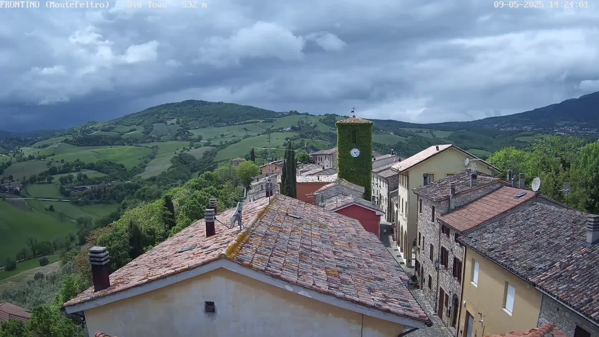 A small village with terracotta roofs nestles in a green valley under a partly cloudy sky.