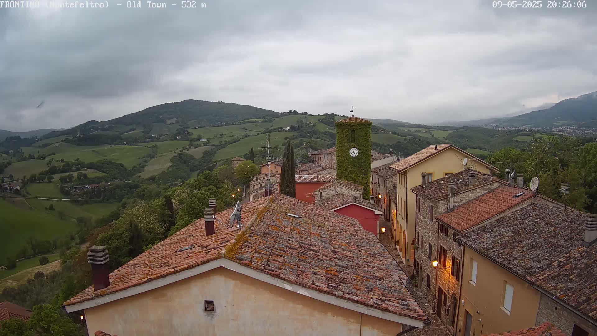 A view from above shows a small town nestled in a valley, with rolling green hills under a cloudy sky.