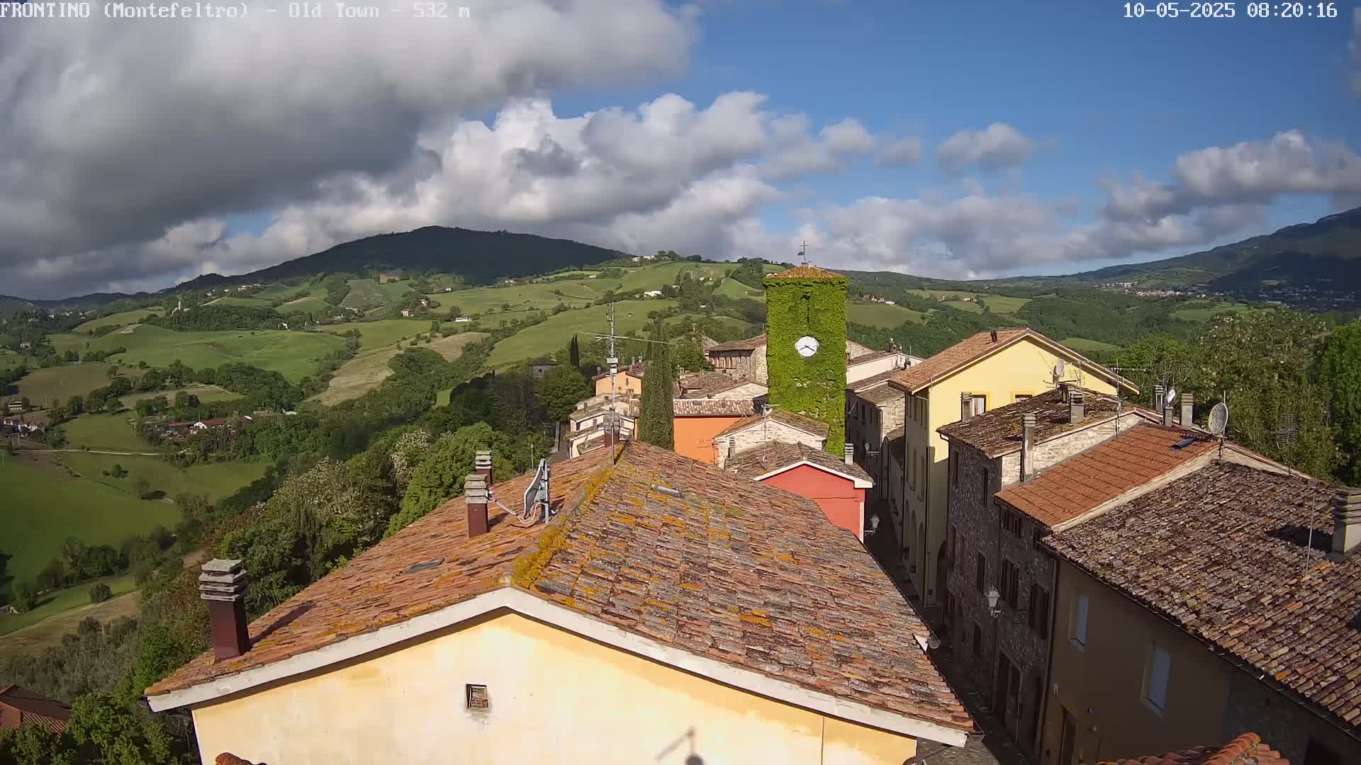 A partly cloudy day overlooks a small village nestled in rolling green hills, with terracotta rooftops and a clock tower covered in ivy.
