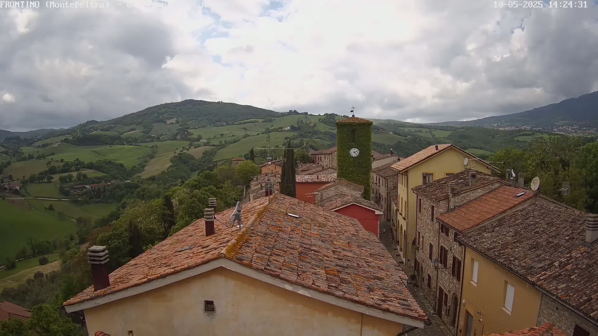 A village of stone buildings with terracotta roofs sits on a hillside overlooking a verdant valley under a partly cloudy sky.