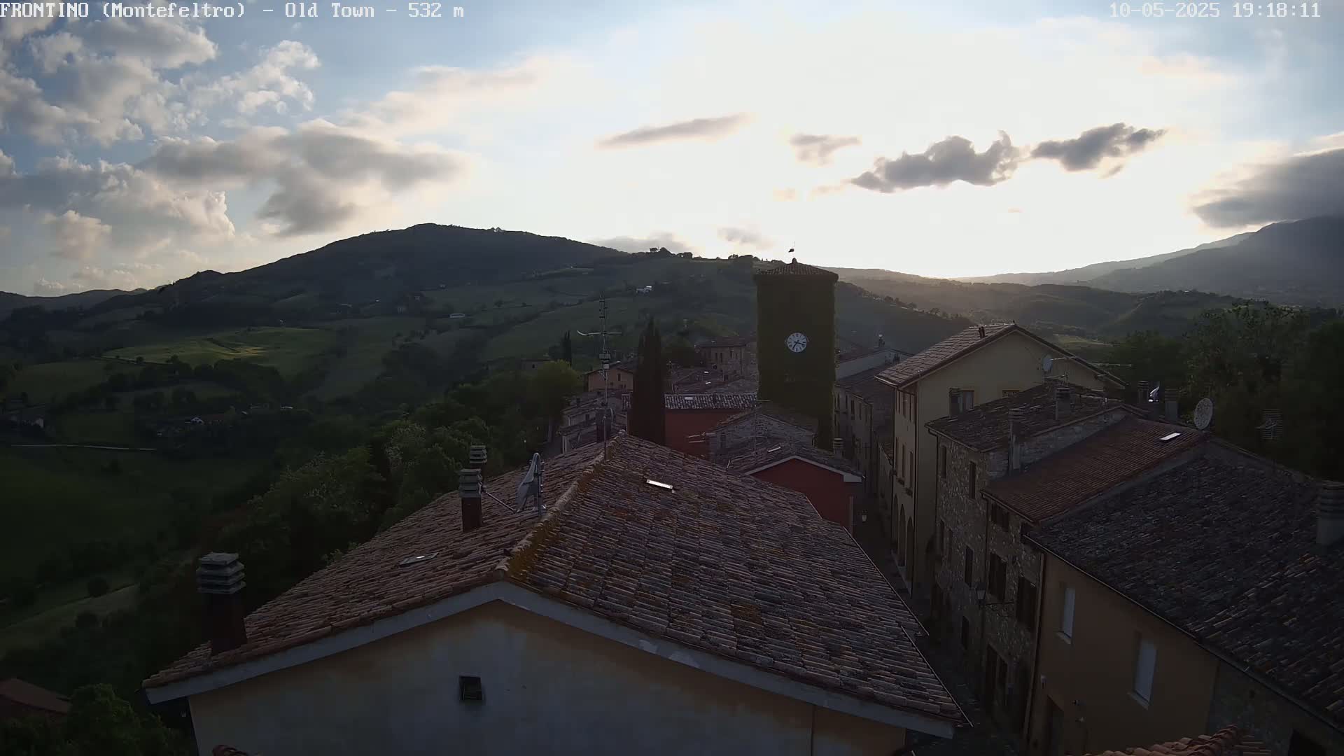 A sunlit hillside town with terracotta roofs nestles in a valley under a partly cloudy sky.