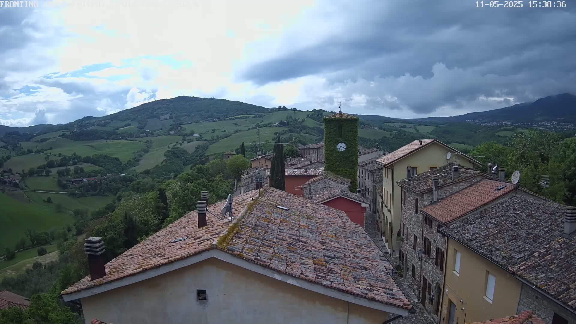 A village of stone and tile-roofed buildings nestled in a green, hilly landscape under a partly cloudy sky.