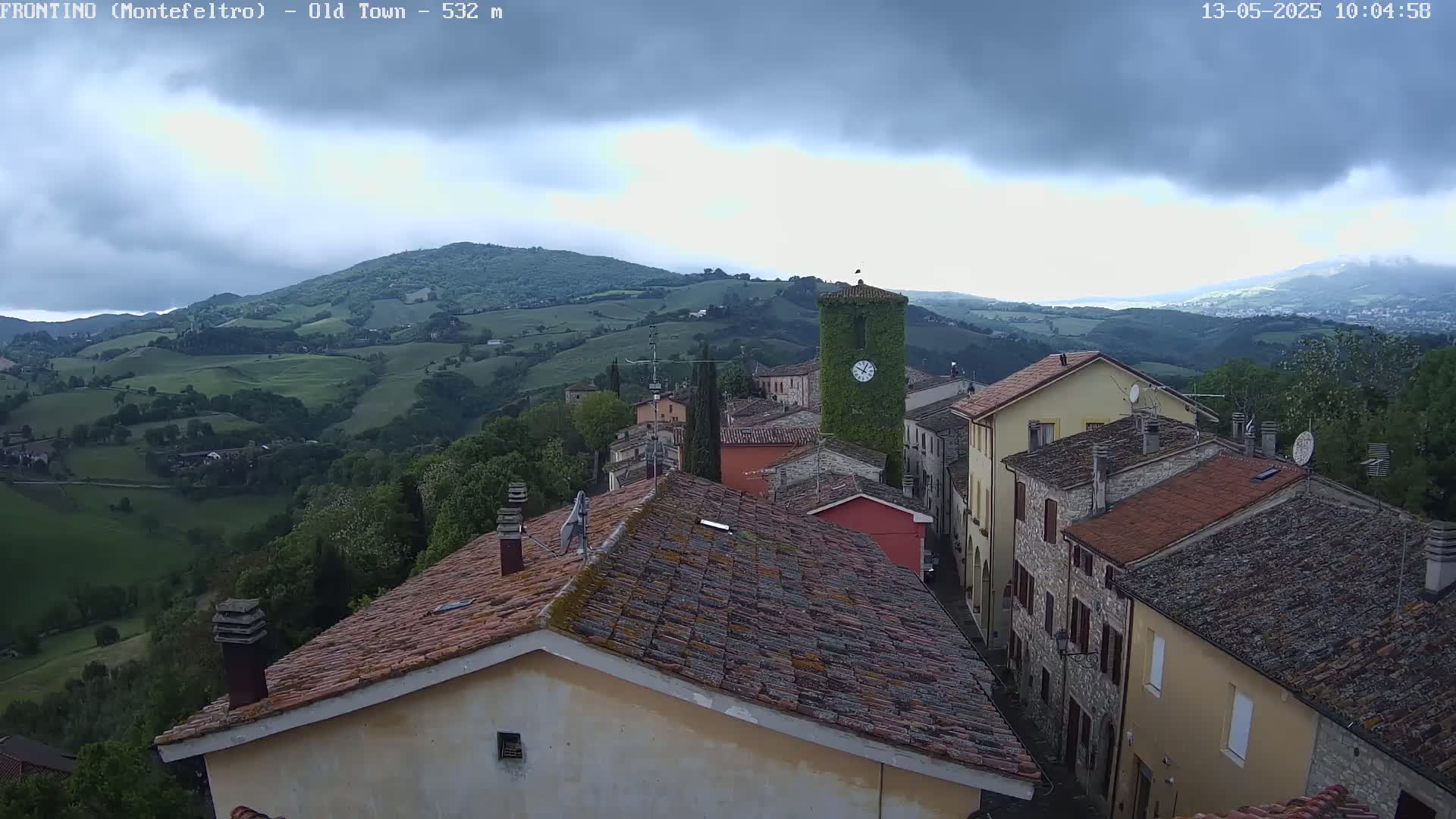 An overcast sky hangs over a hilly, green landscape, overlooking a small village with terracotta rooftops and a clock tower covered in ivy.