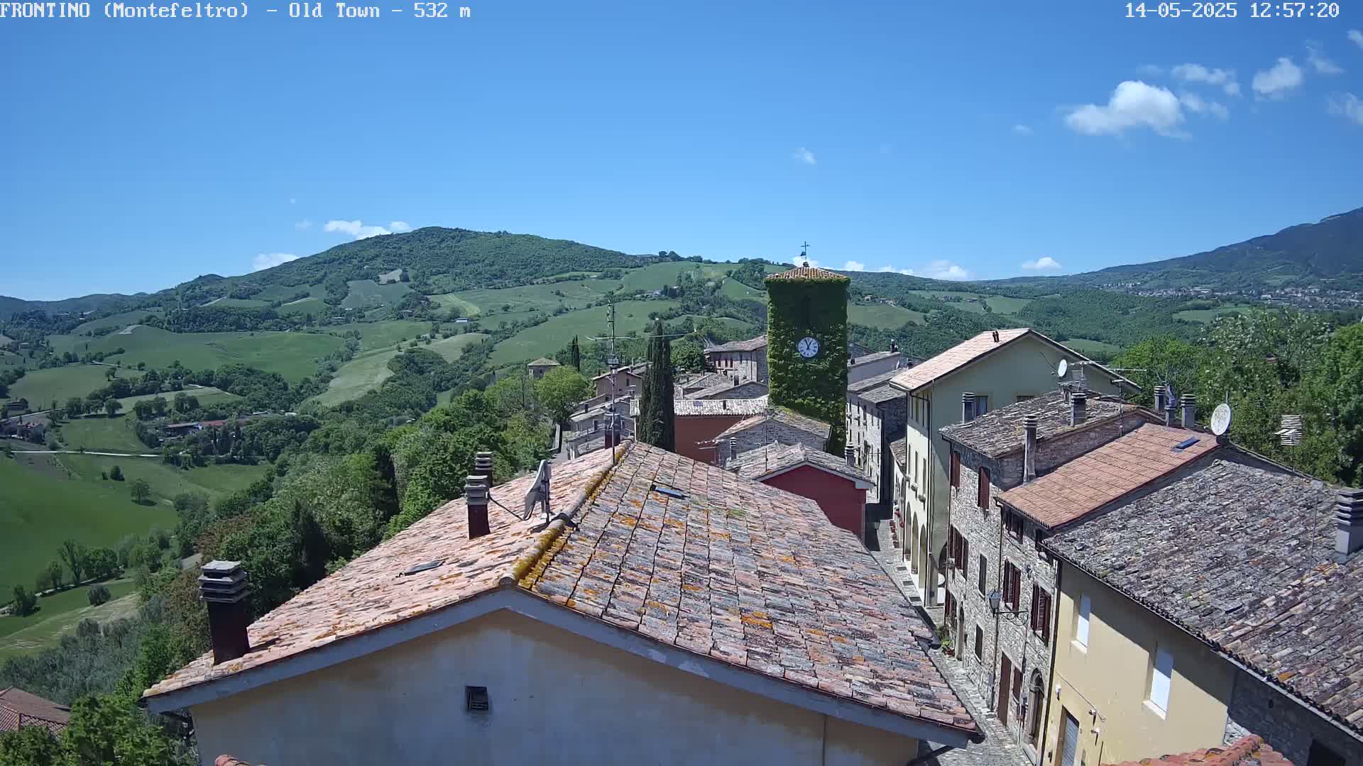 A quaint village nestled in a valley under a mostly clear blue sky, with rolling green hills and mountains in the background.