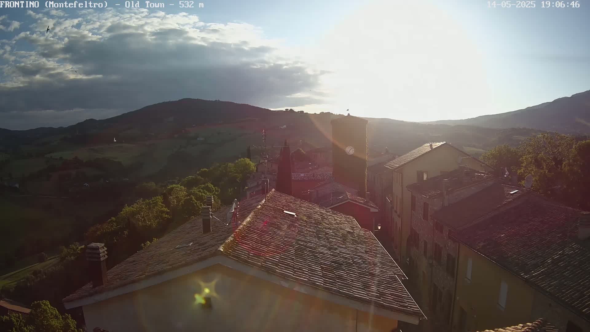 A partly cloudy sunset illuminates a hillside town with stone buildings and terracotta roofs, overlooking a verdant valley.