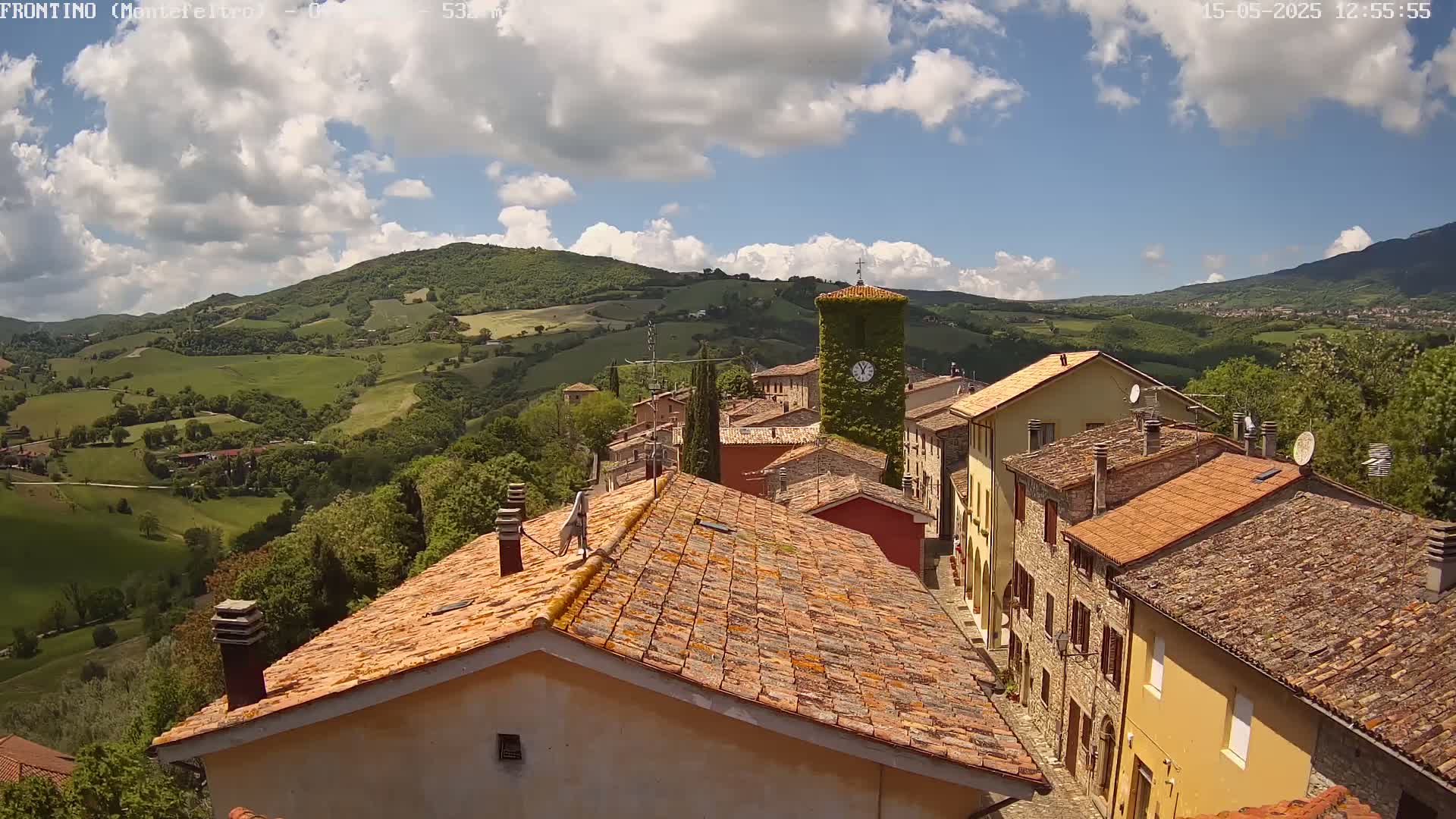 A view from above shows a village of stone buildings with terracotta roofs nestled in a verdant valley under a partly cloudy sky.