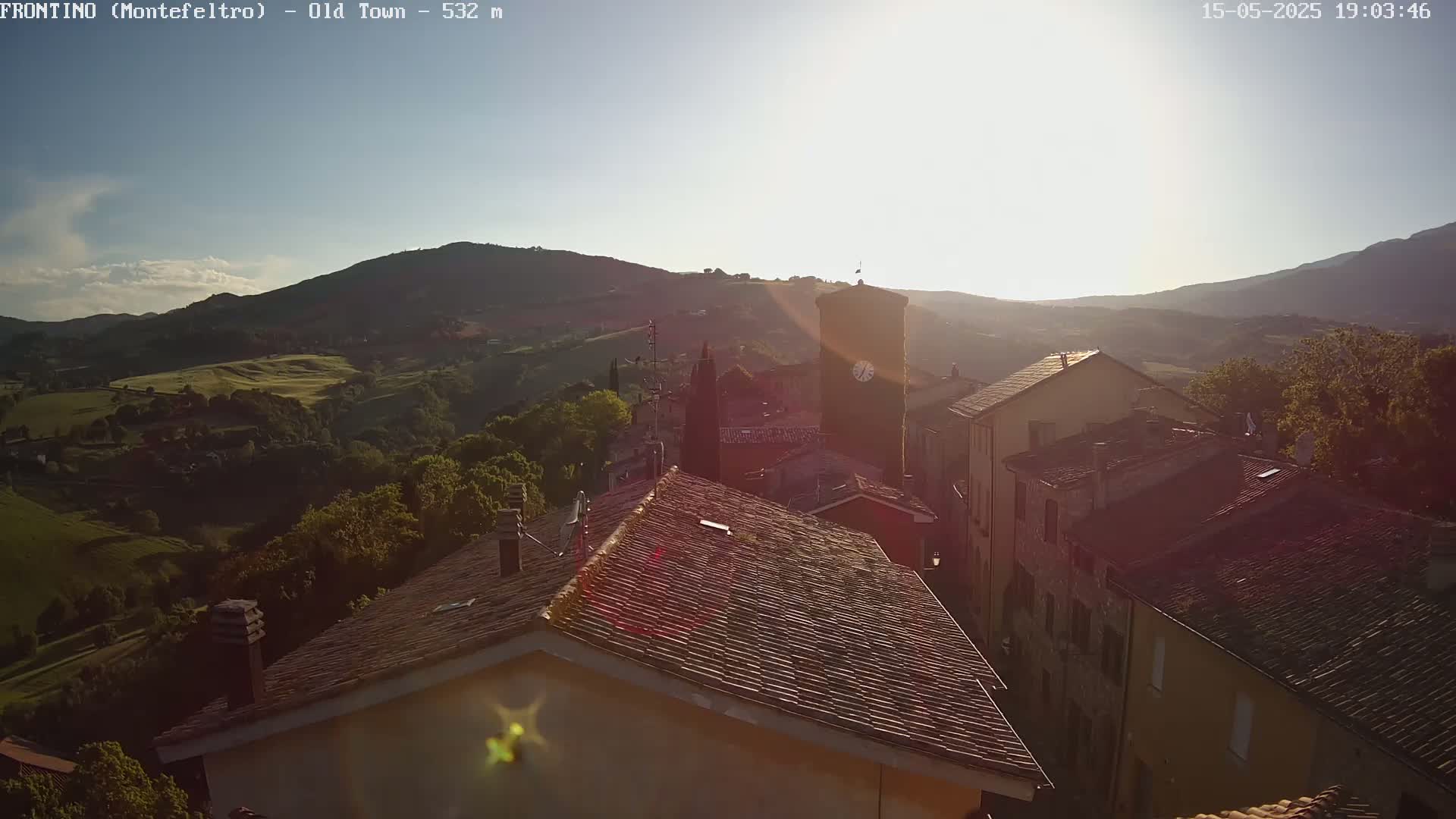 A sunlit view from a rooftop shows a quaint town nestled in rolling green hills under a clear sky.