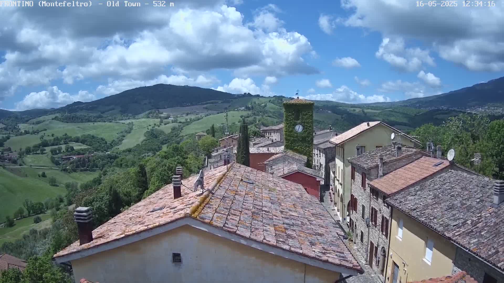 A partly cloudy day overlooks a village nestled in a verdant valley between rolling hills and mountains.
