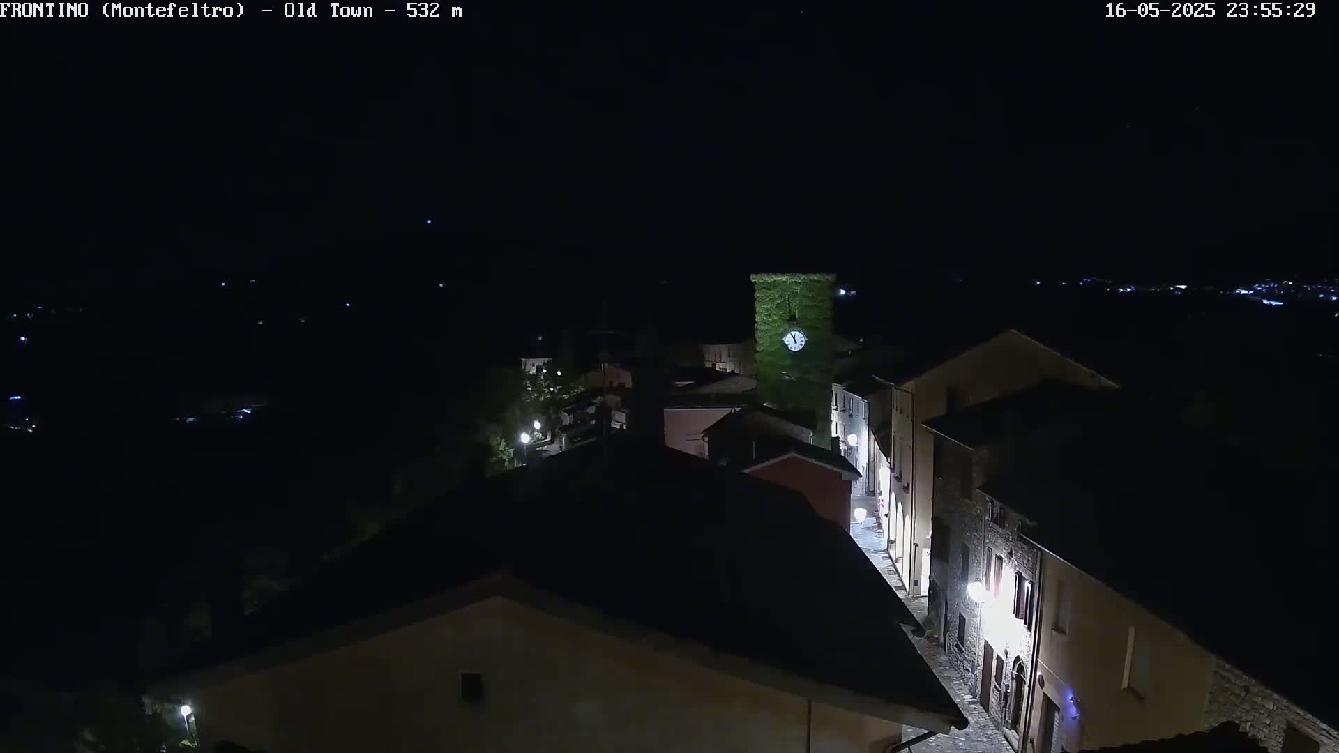A nighttime, high-angle view of a dimly lit, stone village street with buildings and a clock tower under a clear, dark sky.