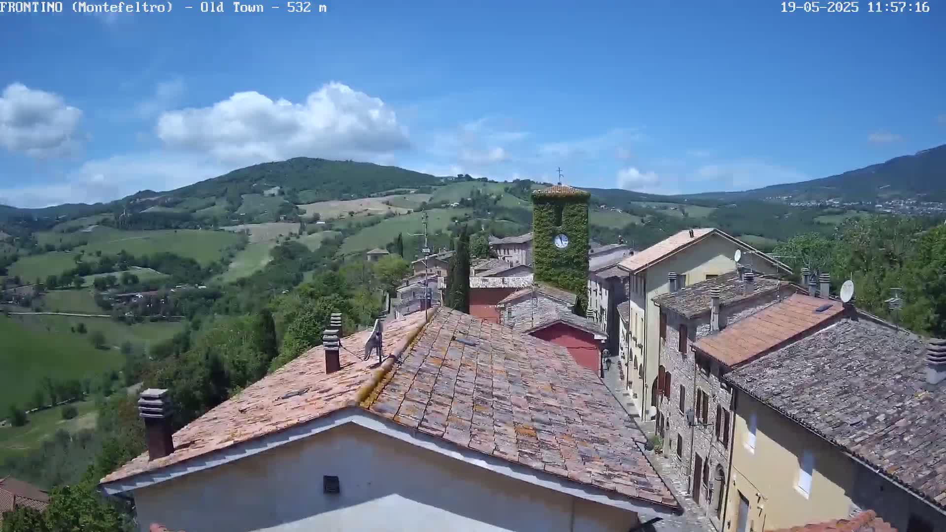 A quaint village nestled in a valley under a mostly sunny sky, with rolling green hills in the background.