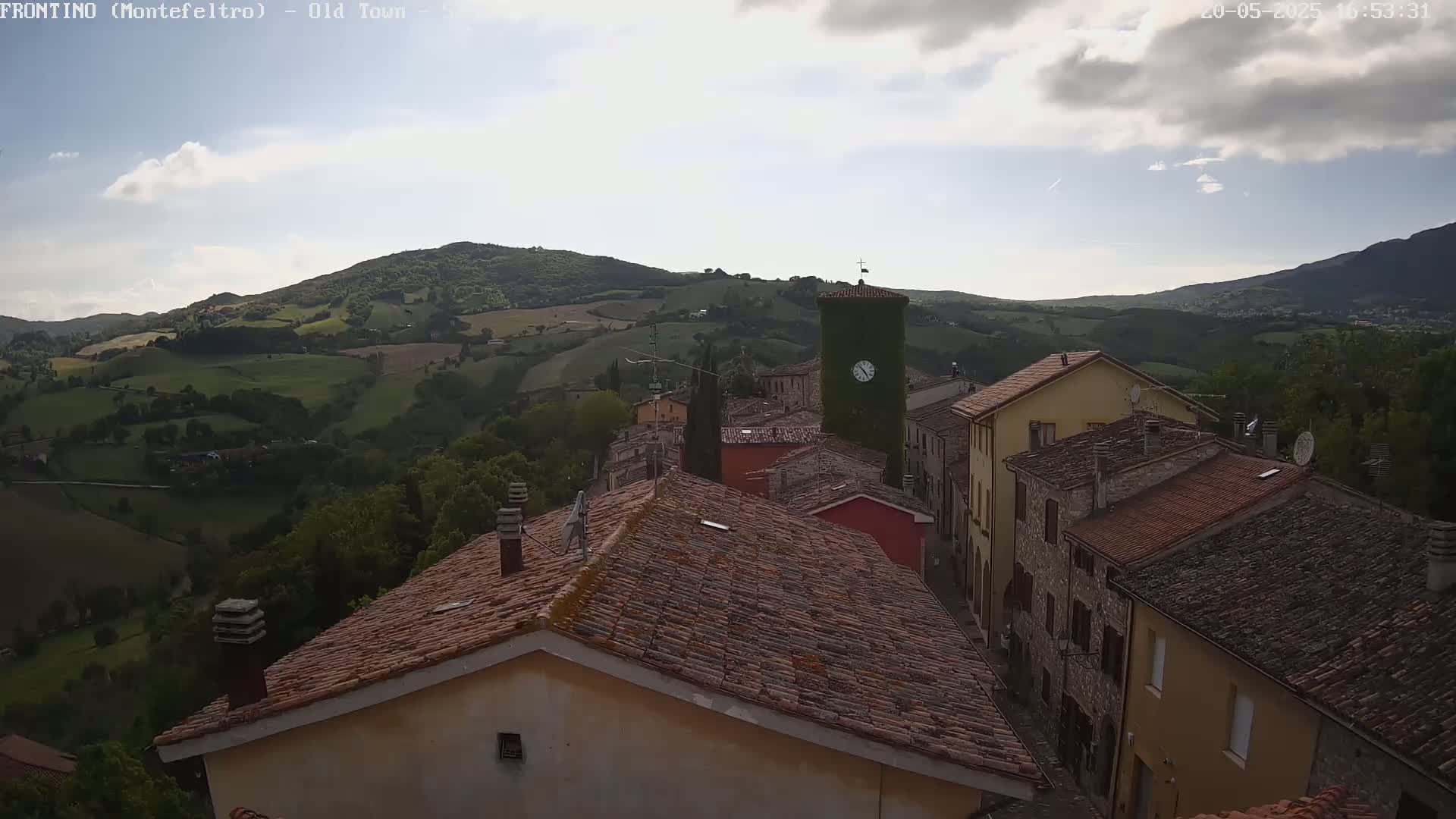 A partly cloudy sky overlooks a terracotta-roofed village nestled in a valley of rolling green hills.