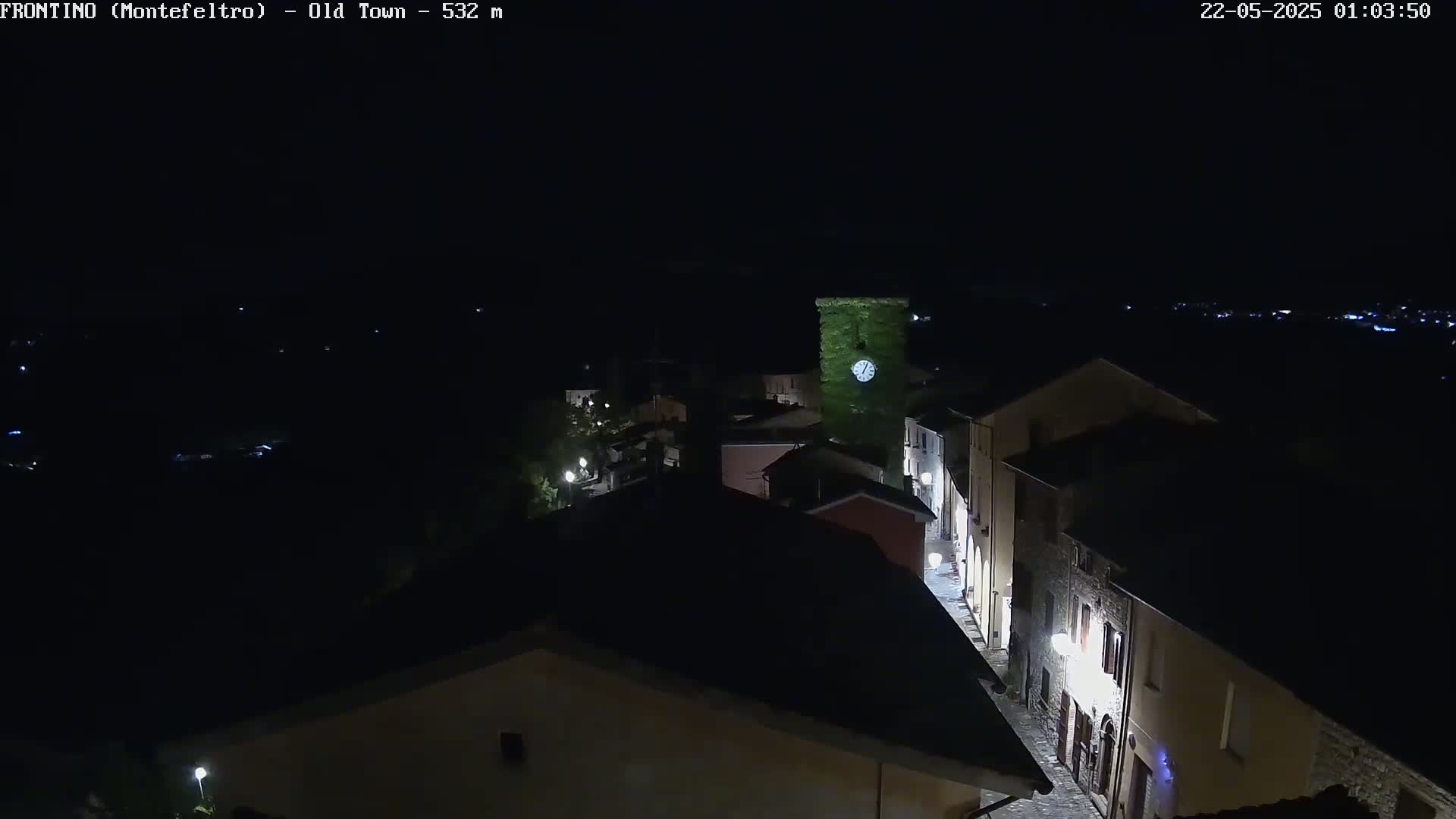 A nighttime, aerial view shows a small town with dimly lit buildings and streets, centered on a clock tower covered in greenery.