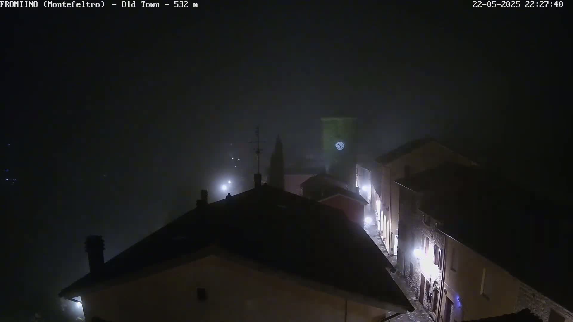 A foggy night scene shows a dimly lit street in a town with buildings, streetlights, and a clock tower visible in the distance.