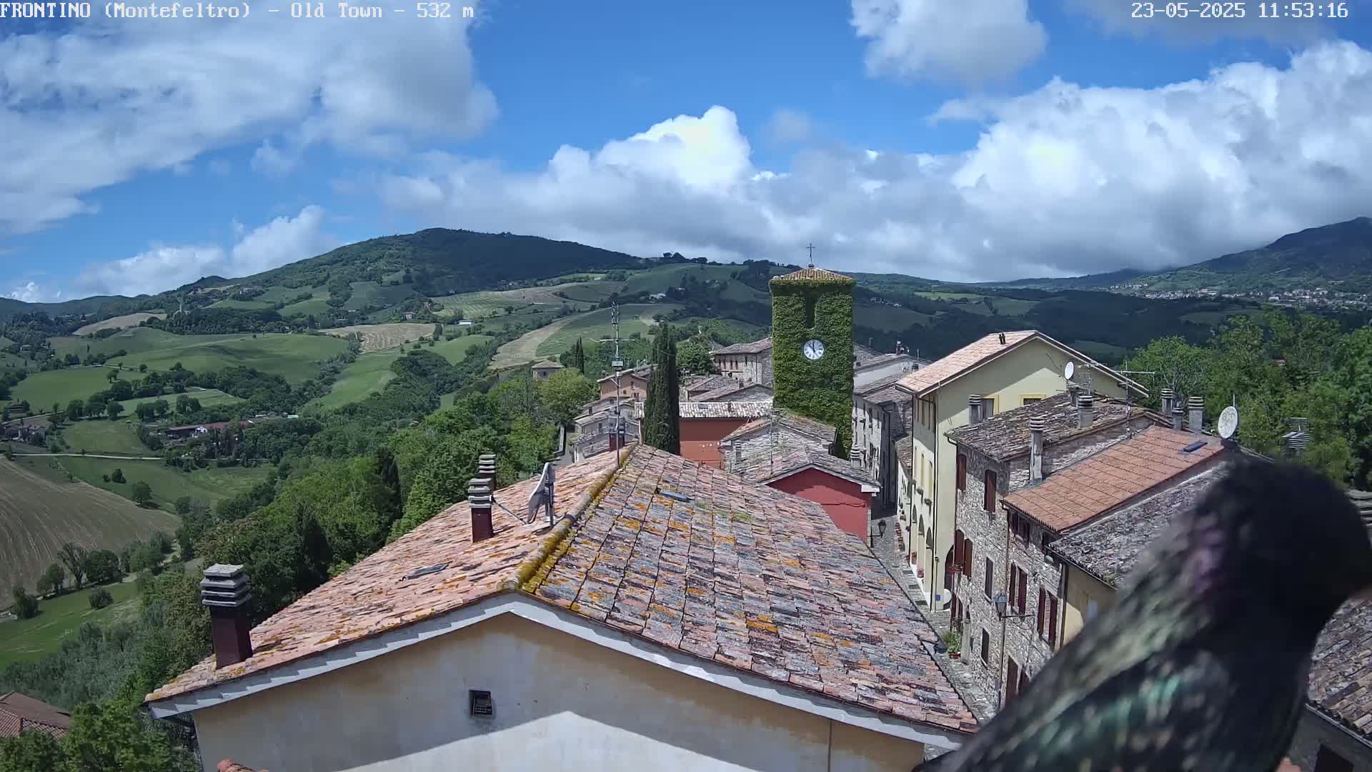 A partly cloudy sky overlooks a small village nestled in a green, hilly landscape.