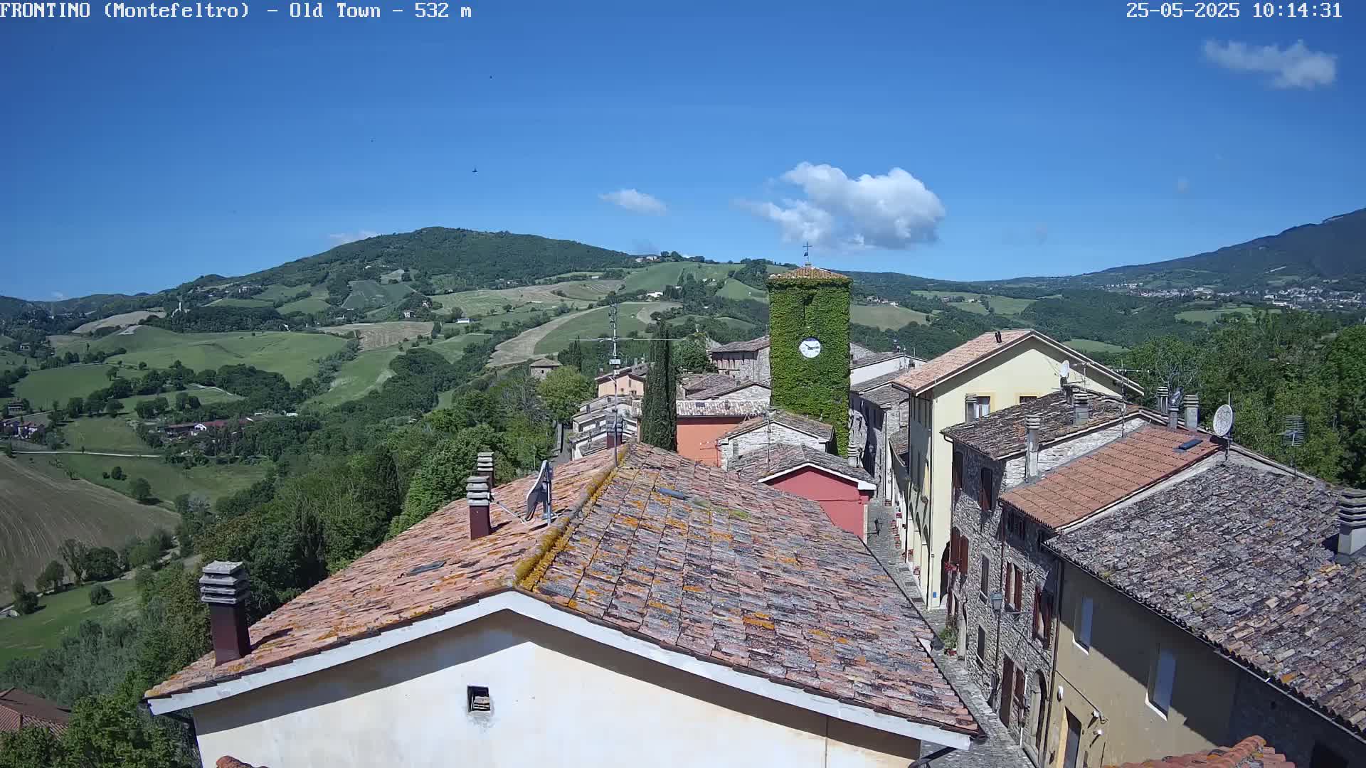 A quaint village with terracotta rooftops nestles in a valley under a mostly sunny sky, surrounded by rolling green hills.
