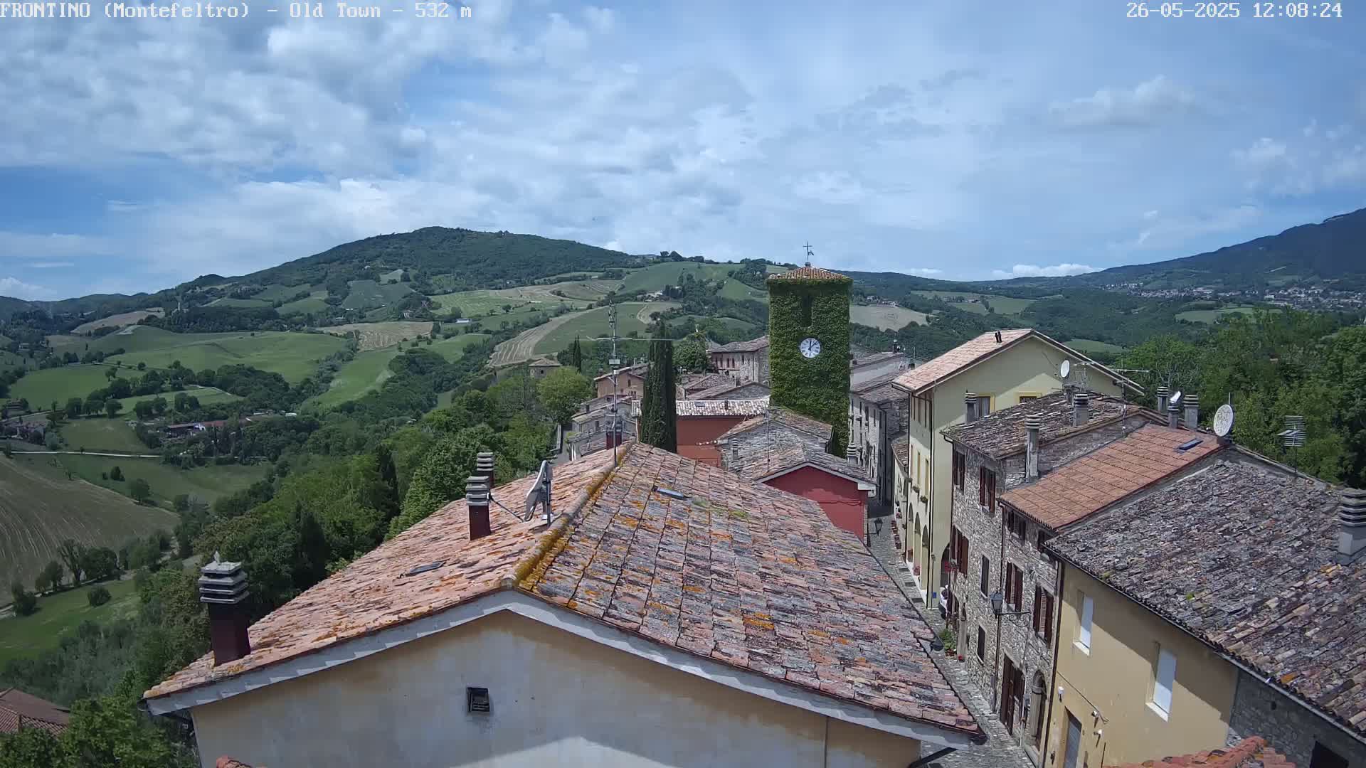 A partly cloudy sky overlooks a village of terracotta-roofed buildings nestled in a verdant valley.