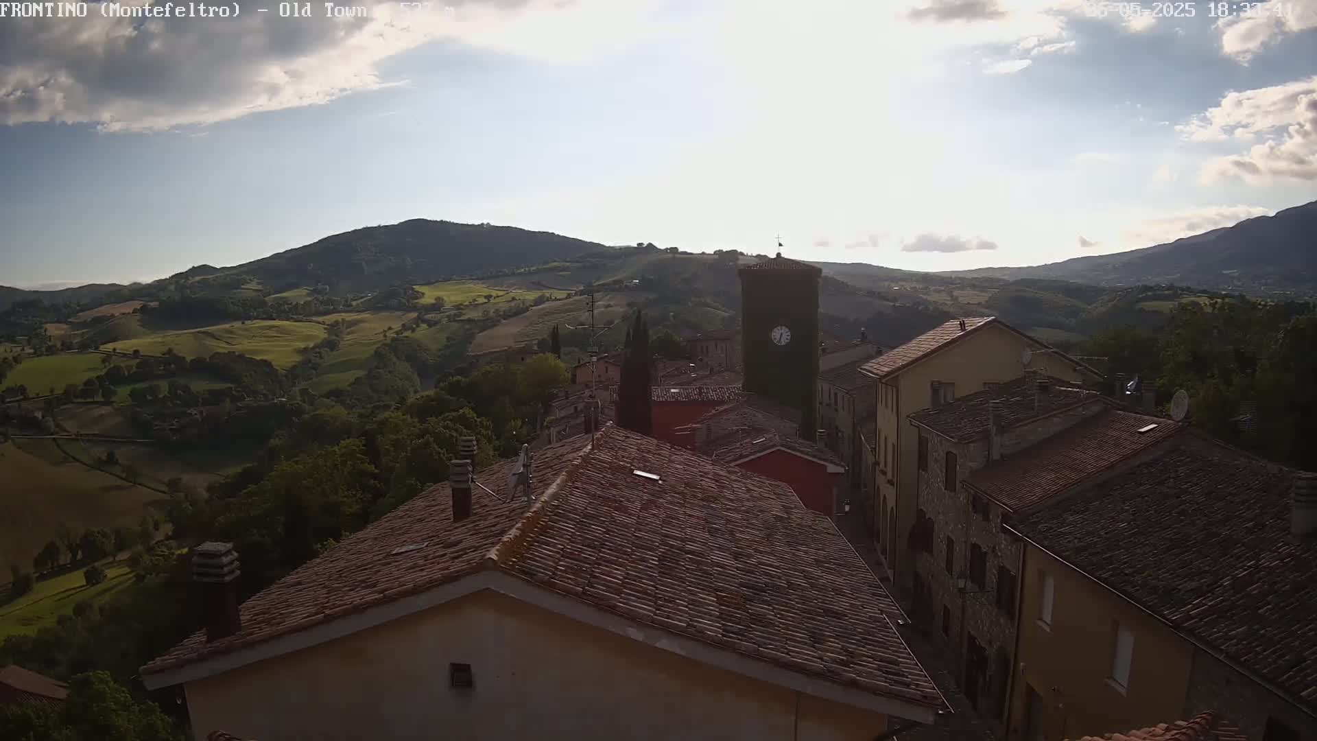 A partly sunny view overlooks a village of red-tiled roofs nestled in a valley surrounded by rolling green hills.