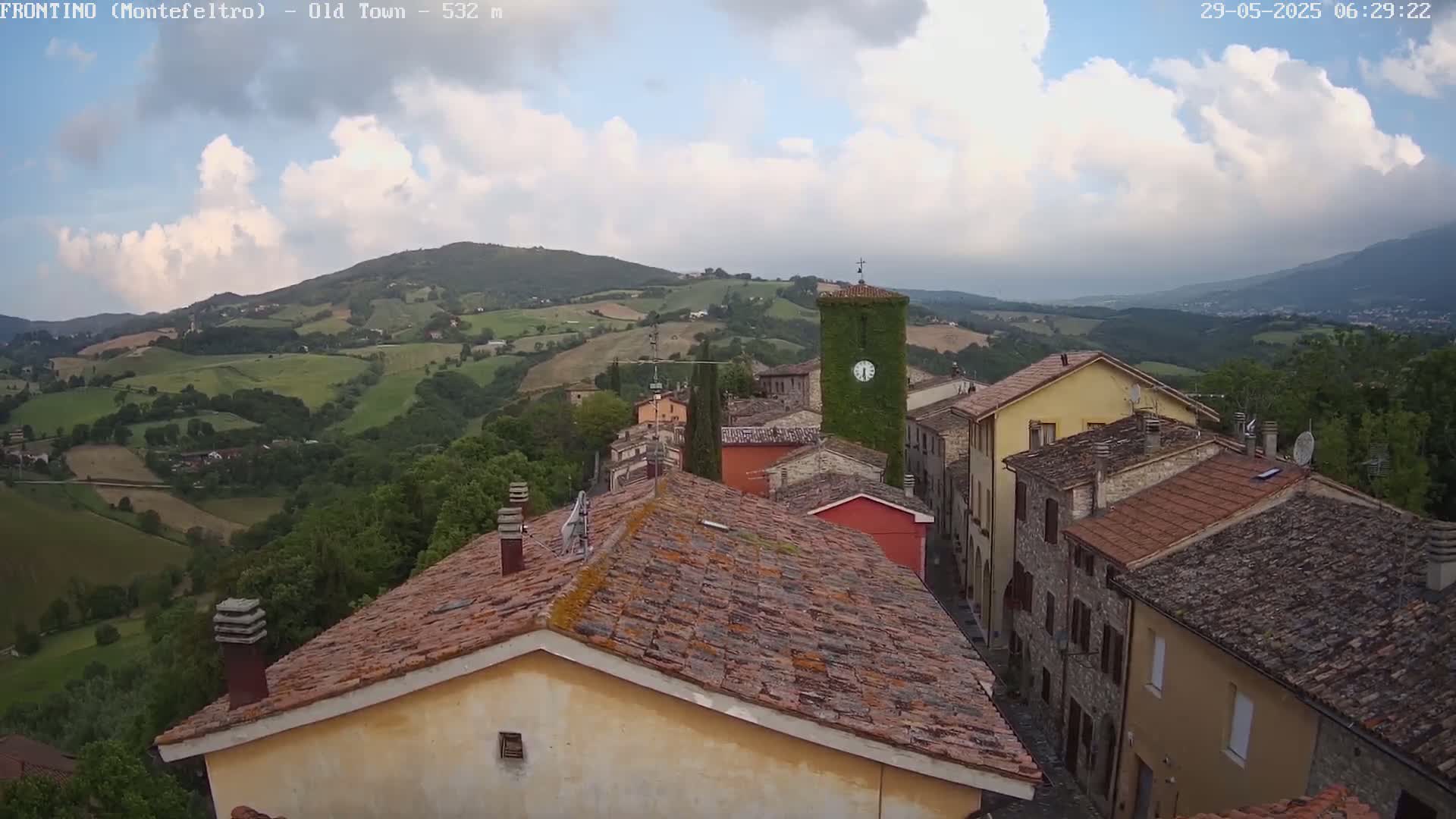 A partly cloudy sky overlooks a quaint village nestled in a rolling green hillside.