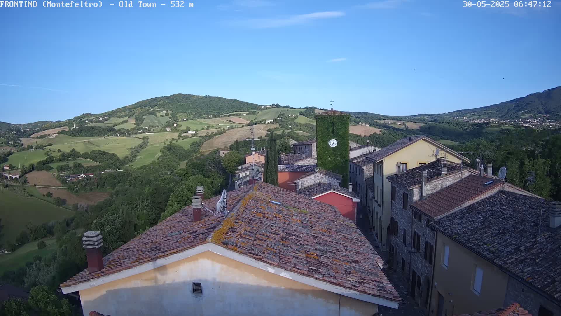 An aerial view of a small village nestled in a valley on a clear, sunny day, with rolling green hills in the background.