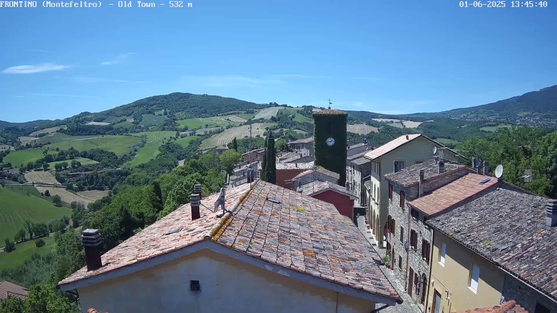 A view of a small town nestled in a hilly landscape under a clear blue sky shows terracotta-tiled rooftops and a clock tower, with green rolling hills extending into the distance.