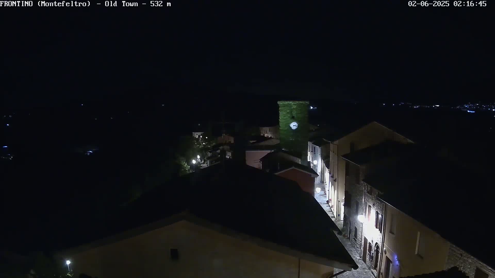A nighttime, aerial view shows a small, dimly lit town with a clock tower covered in greenery.