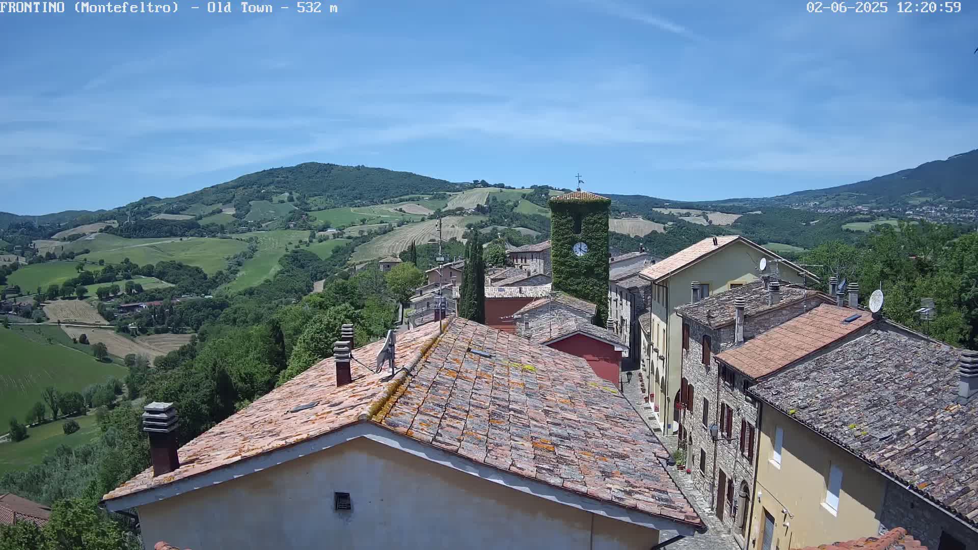 A picturesque village with terracotta rooftops nestles in a valley under a clear blue sky, surrounded by rolling green hills.