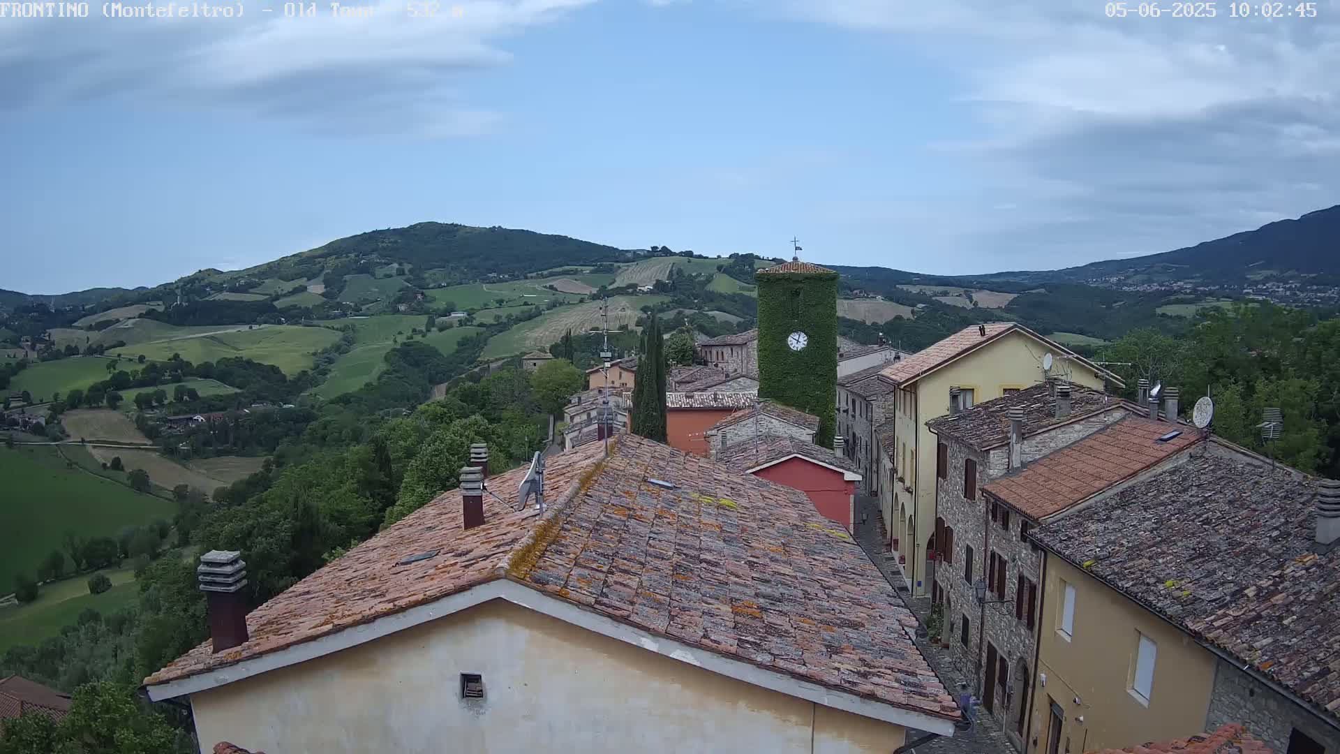 A view from a rooftop shows a village nestled in a valley, with rolling green hills and a partly cloudy sky.
