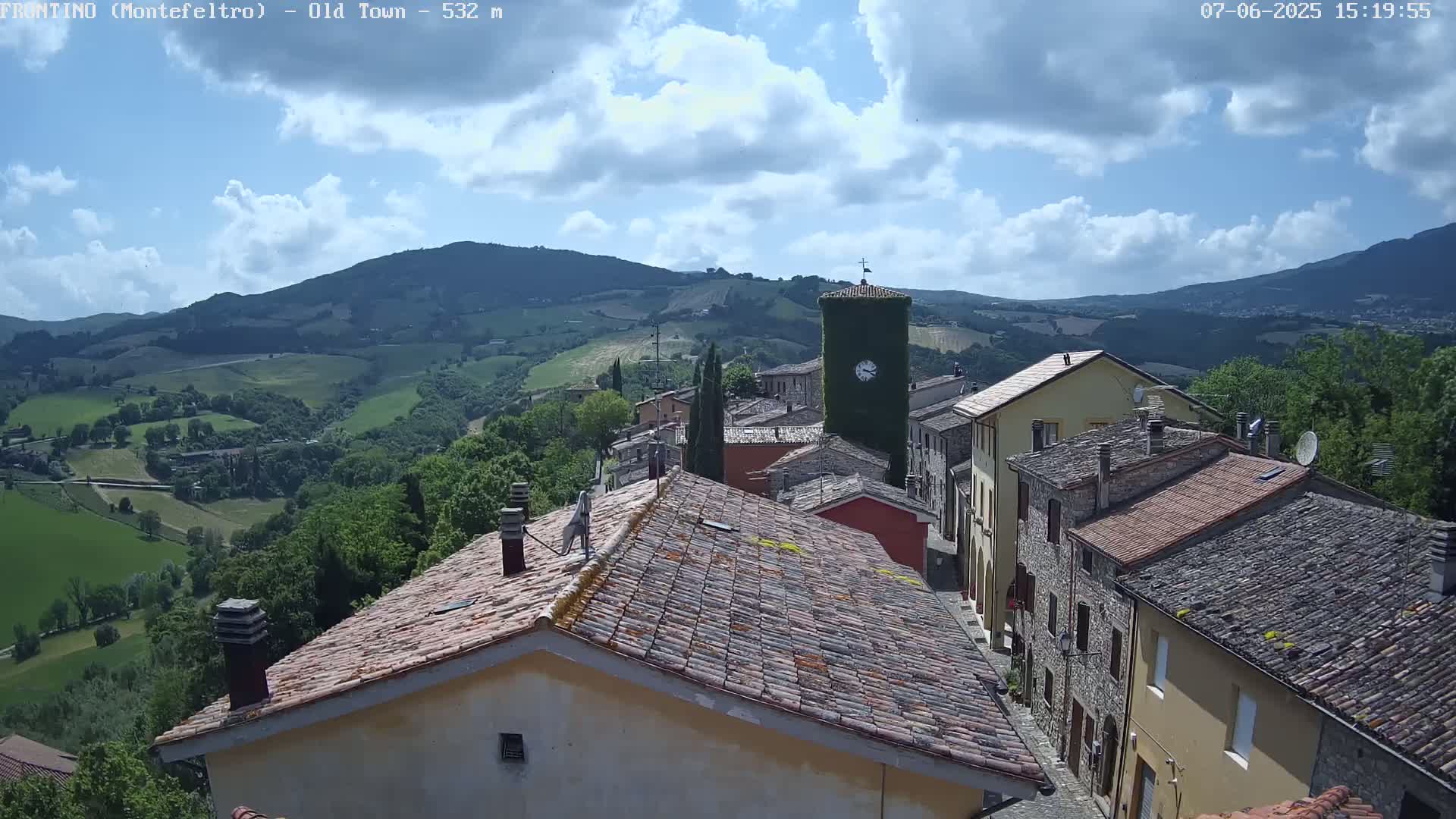 A partly cloudy day overlooks a small village nestled in a valley between rolling green hills, with a clock tower visible in the center.