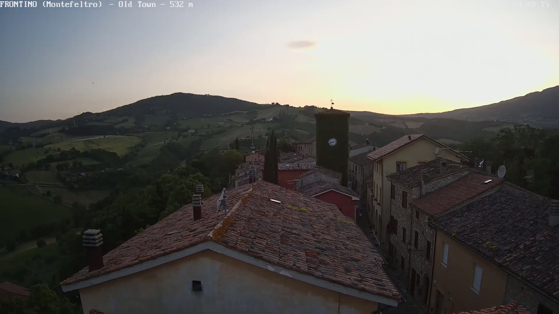 A hilltop village with terracotta roofs is silhouetted against a setting sun over rolling green hills.