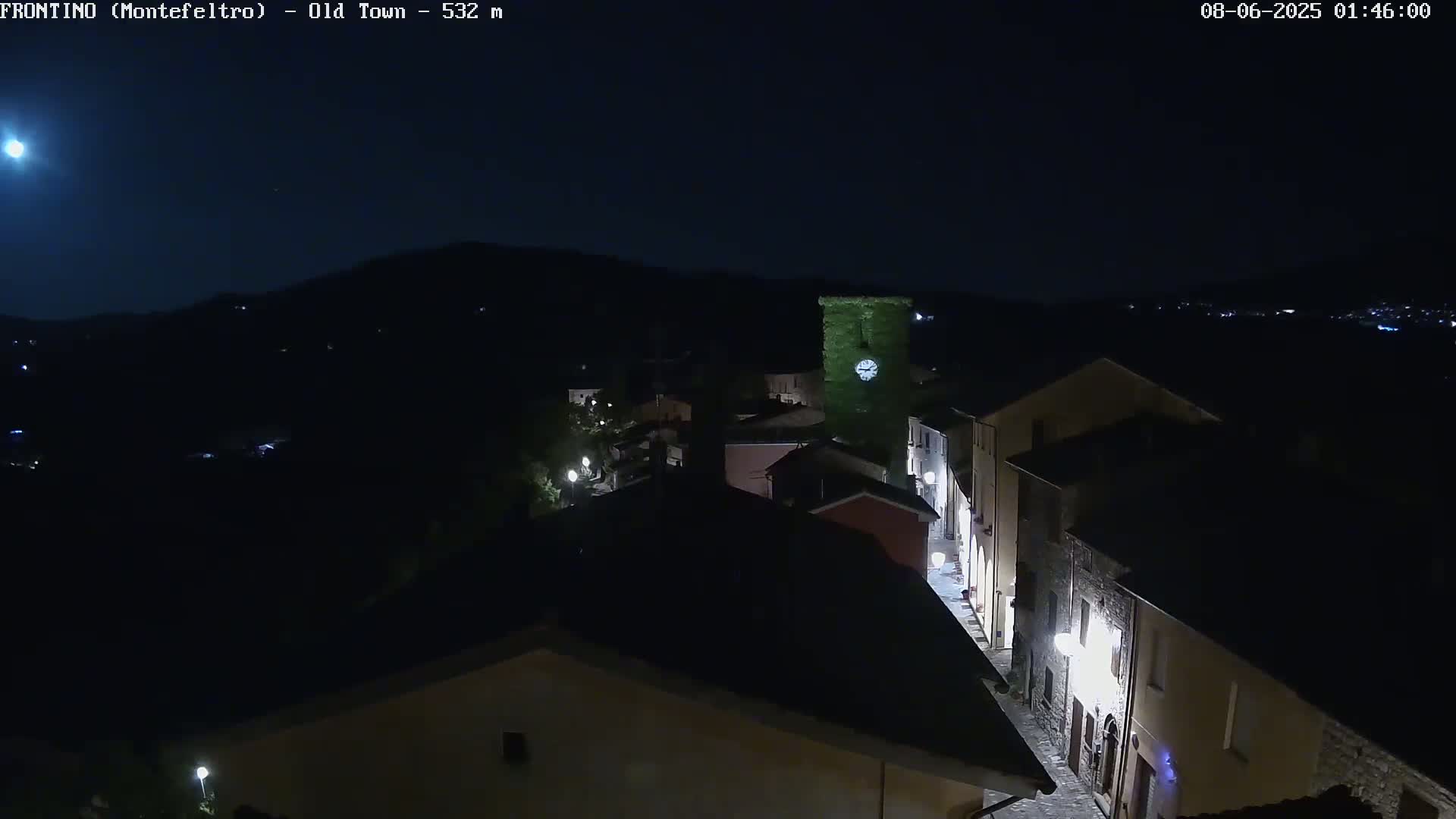 A nighttime, high-angle view shows a small town nestled in a dark, hilly landscape under a clear, moonlit sky.