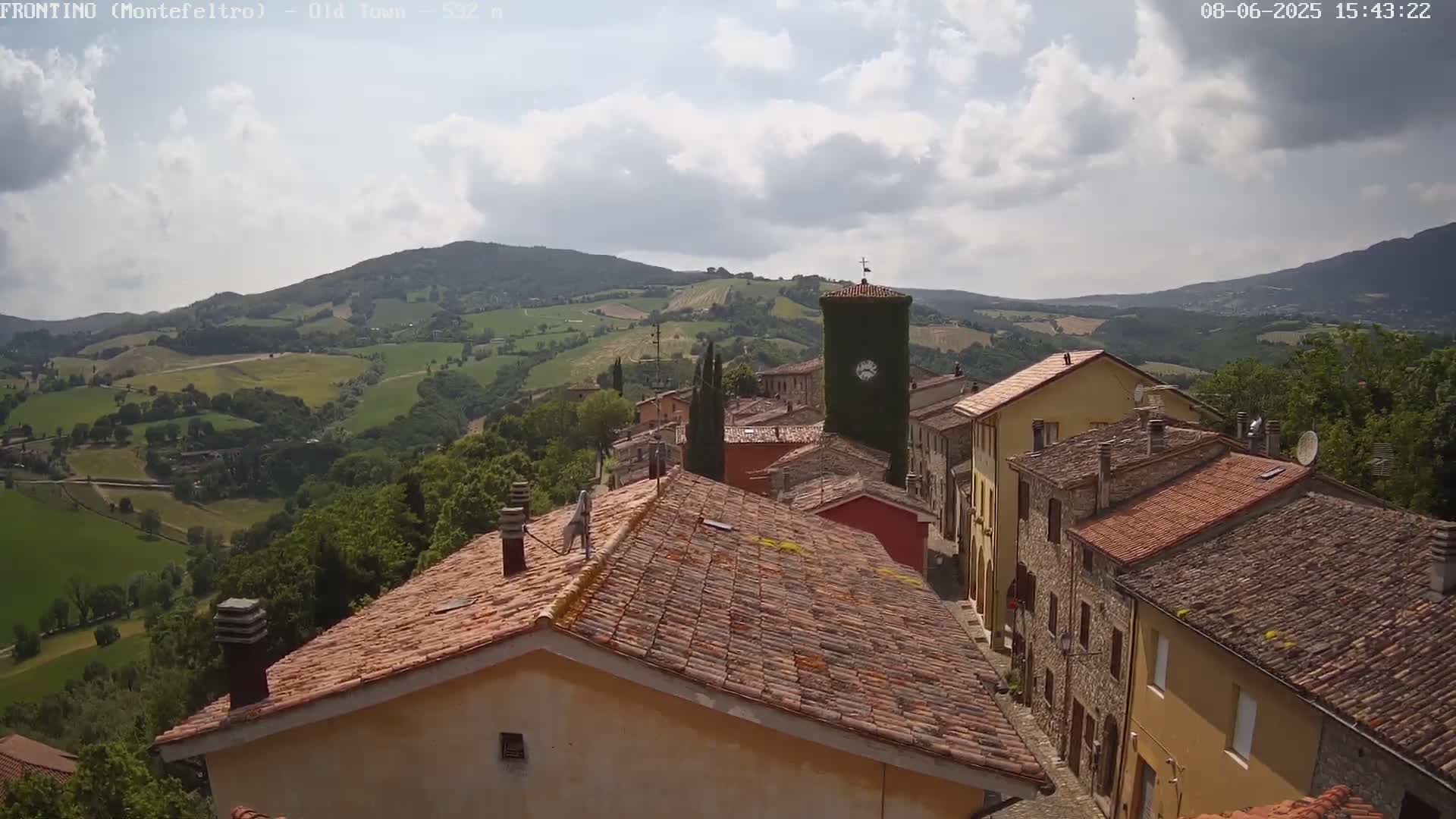 A partly cloudy day reveals a quaint village nestled in a hilly, verdant landscape, with terracotta rooftops dominating the foreground.