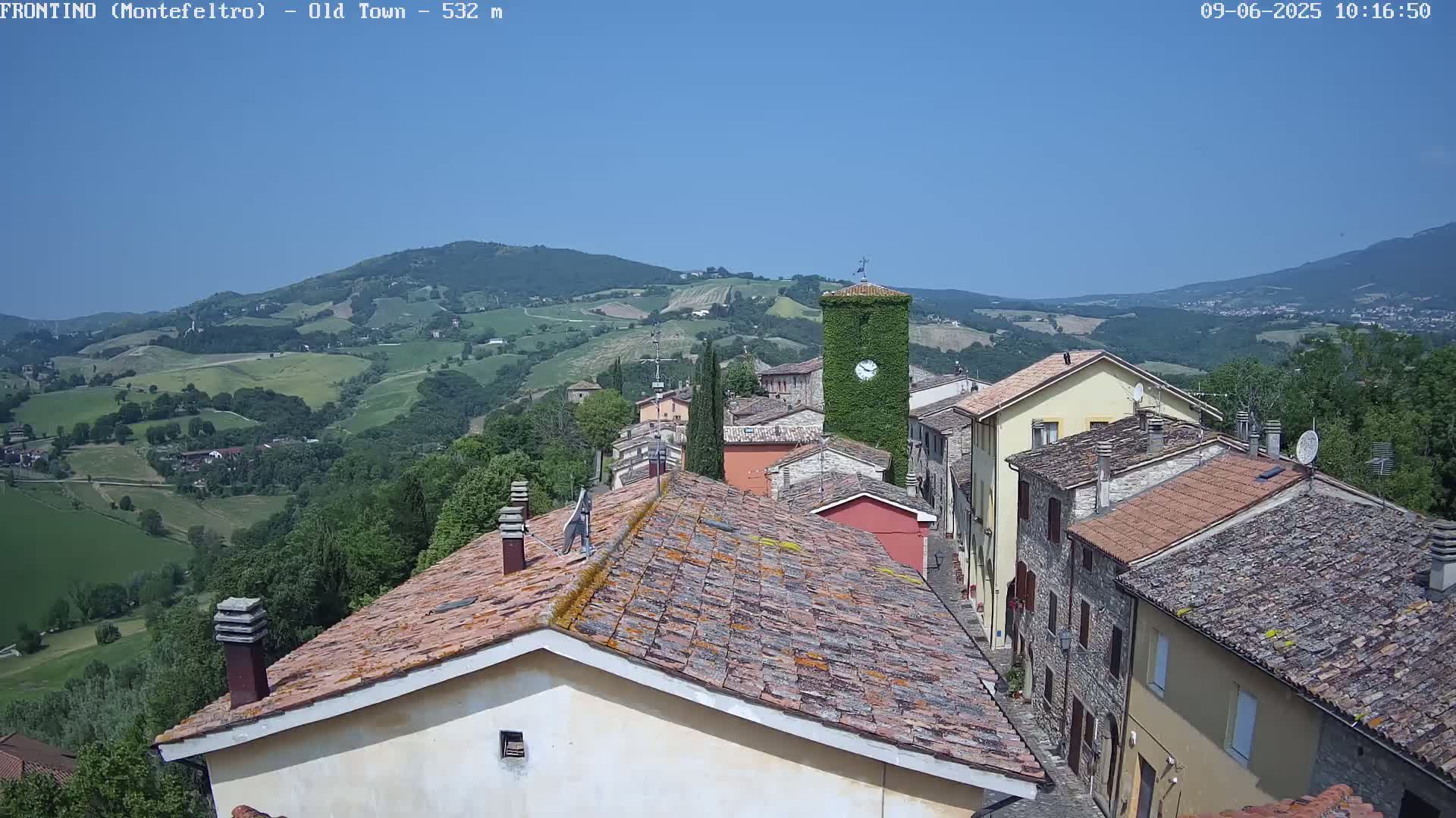 A quaint village nestled in a verdant valley under a clear blue sky, features terracotta rooftops and a clock tower covered in ivy.