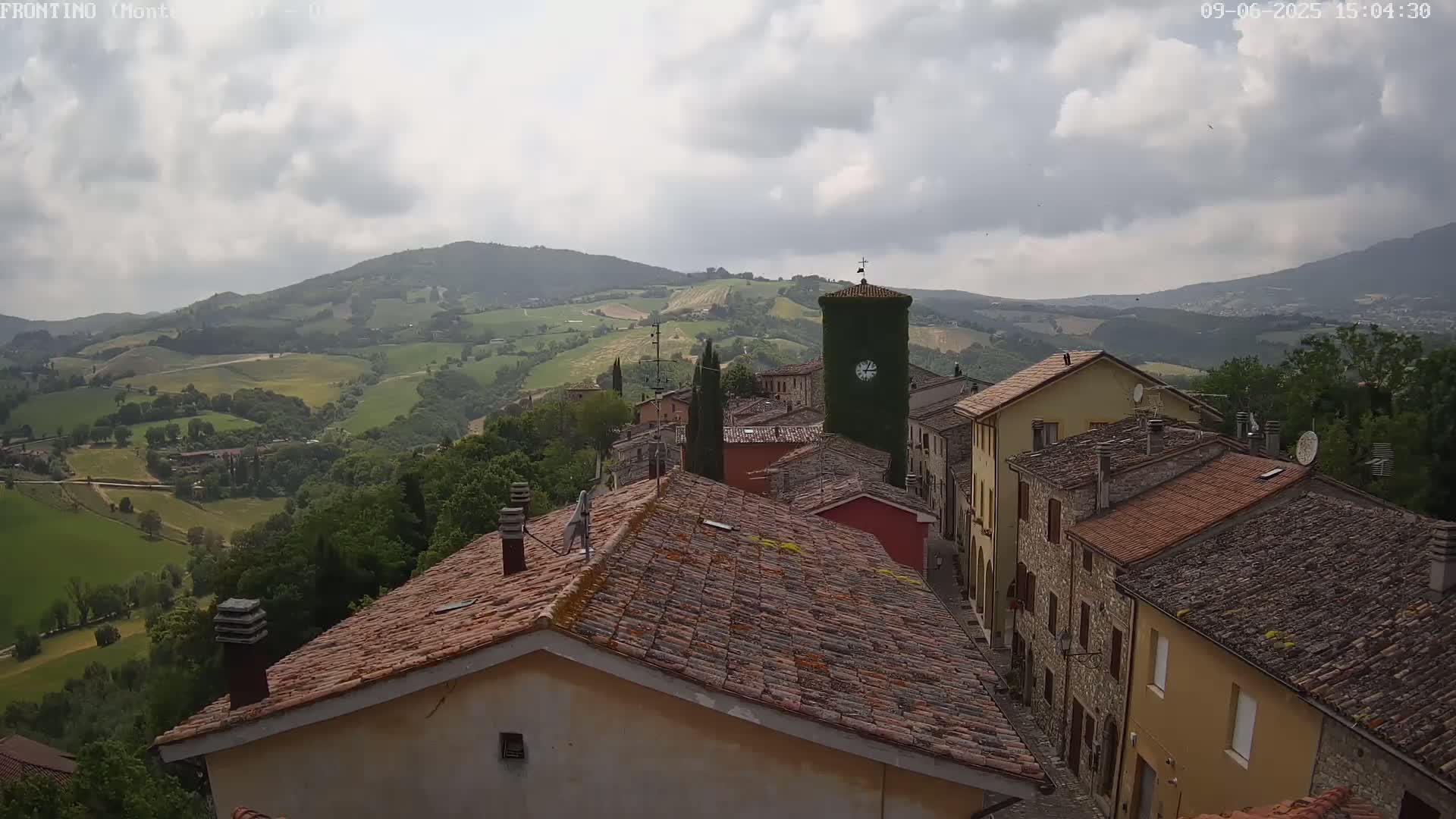 A hilltop village with terracotta roofs overlooks a verdant valley under a cloudy sky.