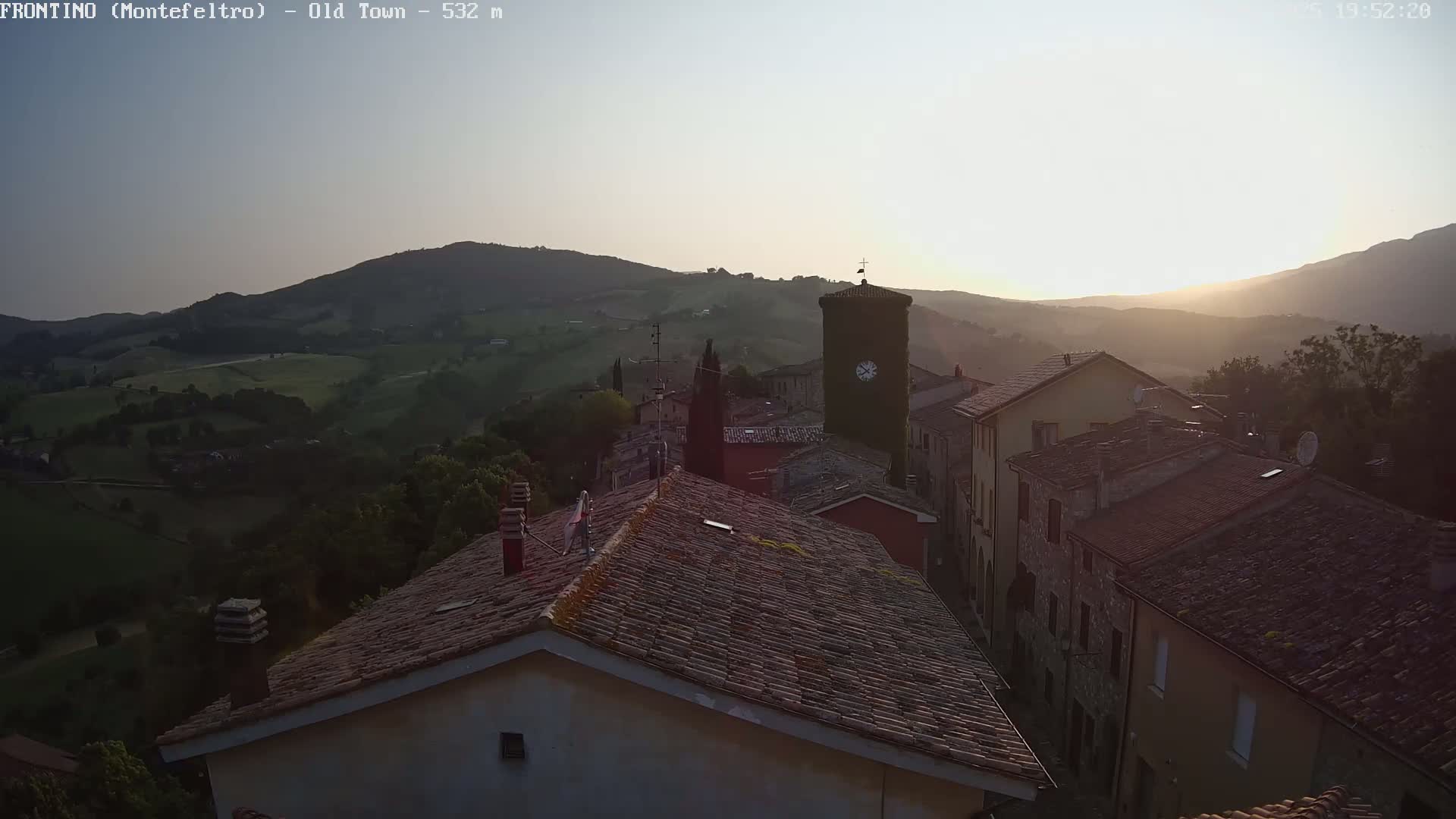 A sunlit view of a small town nestled in rolling hills, showing rooftops of stone and tile buildings with a clock tower at the center.