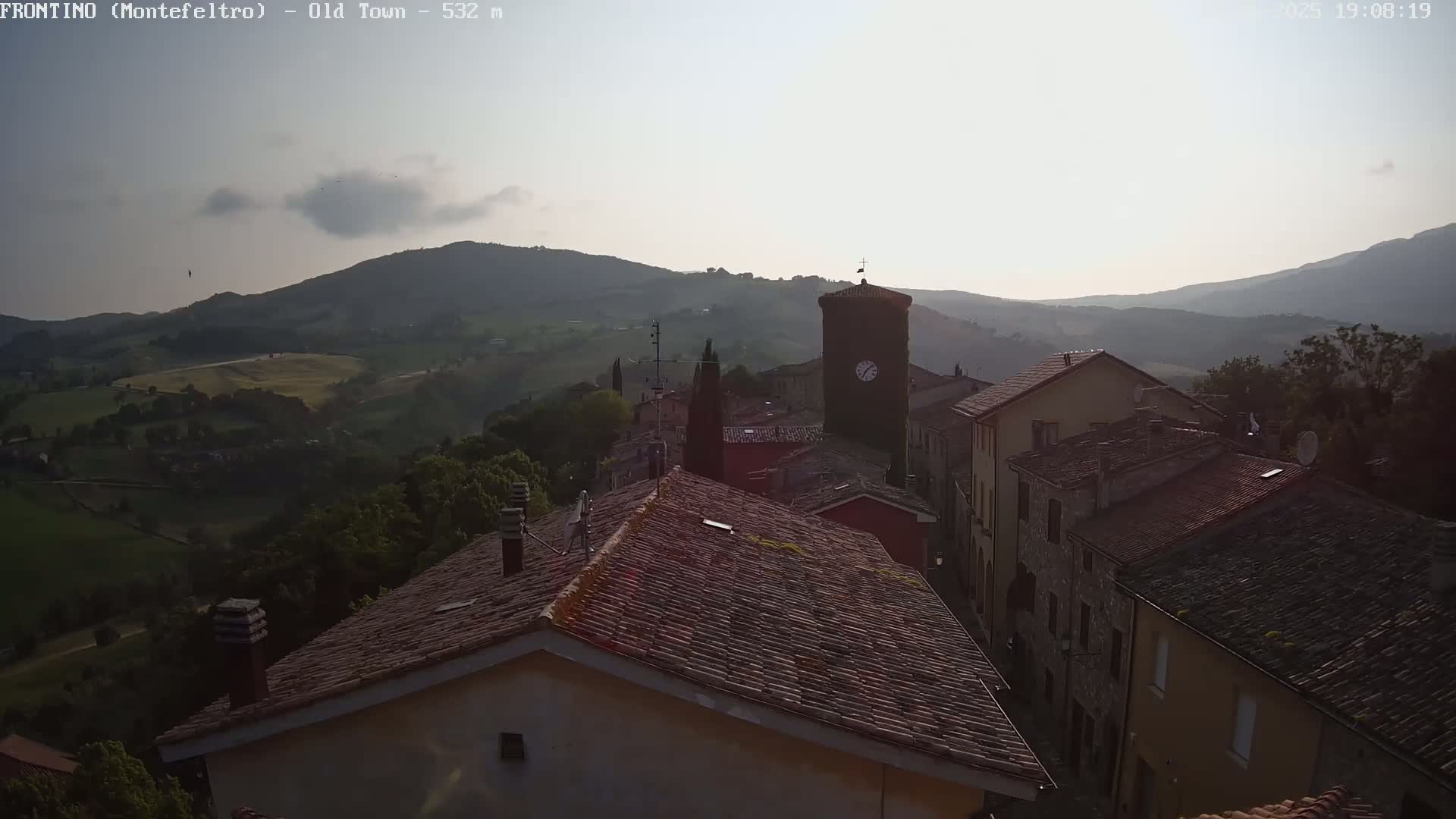 A view from a rooftop shows a quaint town nestled in rolling hills under a mostly clear, light sky.