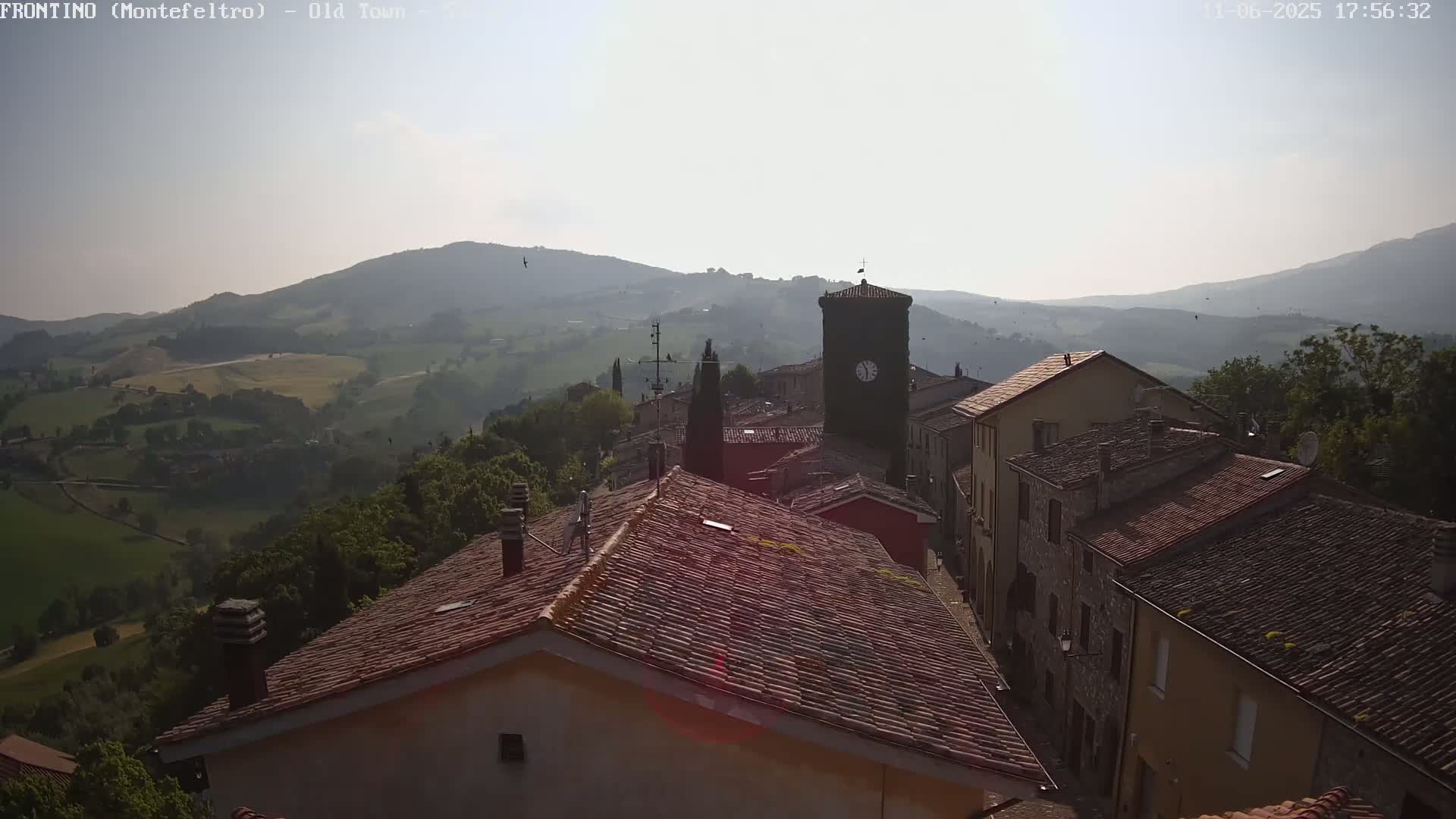 A hazy, sunny day reveals a hillside village with terracotta roofs and a clock tower, overlooking a verdant valley.