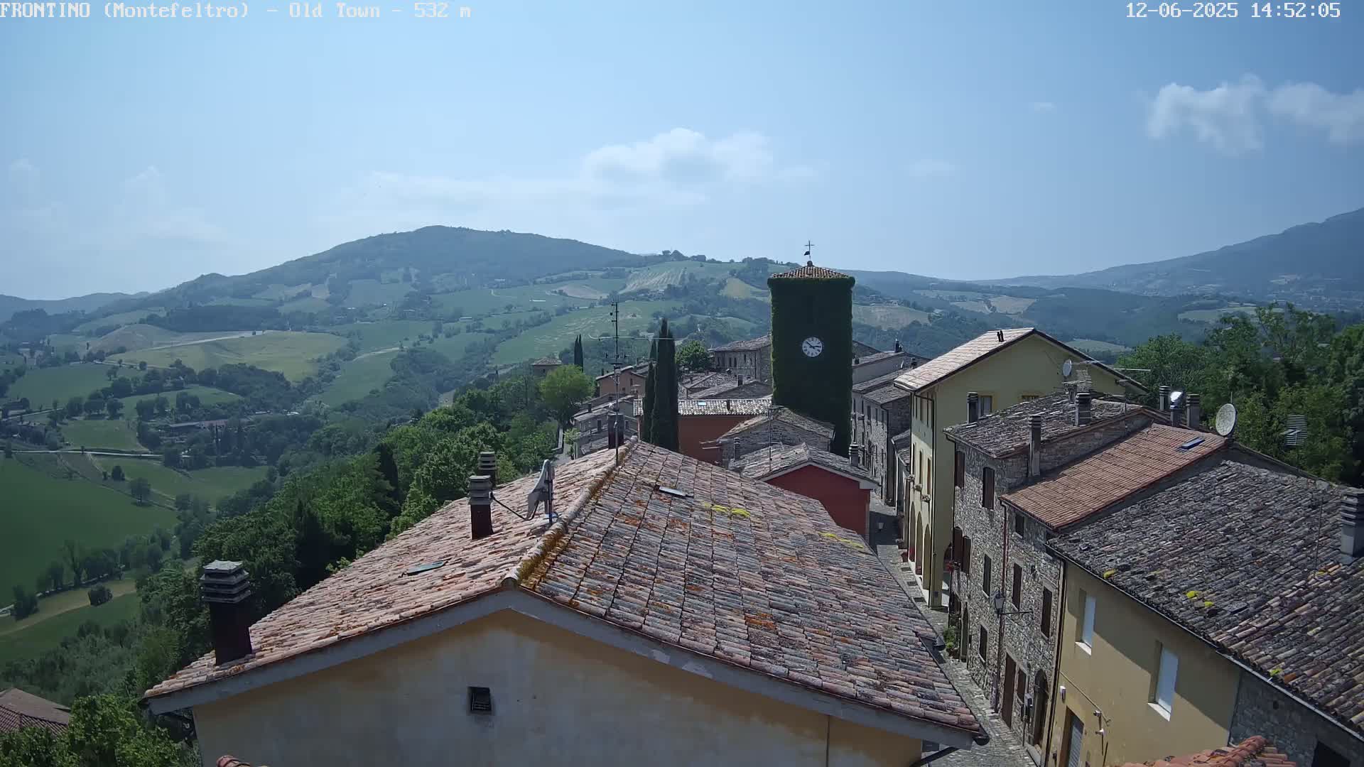 A quaint village with terracotta roofs nestles in a rolling green hillside under a mostly clear blue sky.