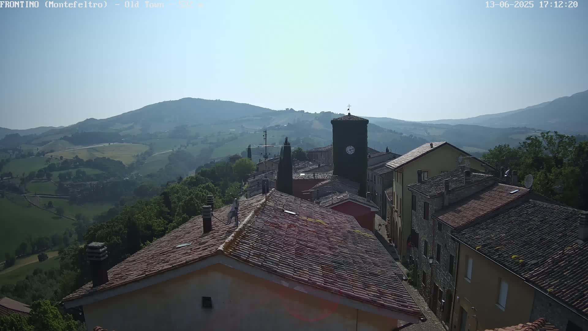 A clock tower stands in the center of a stone village nestled in a valley, under a clear, bright sky.