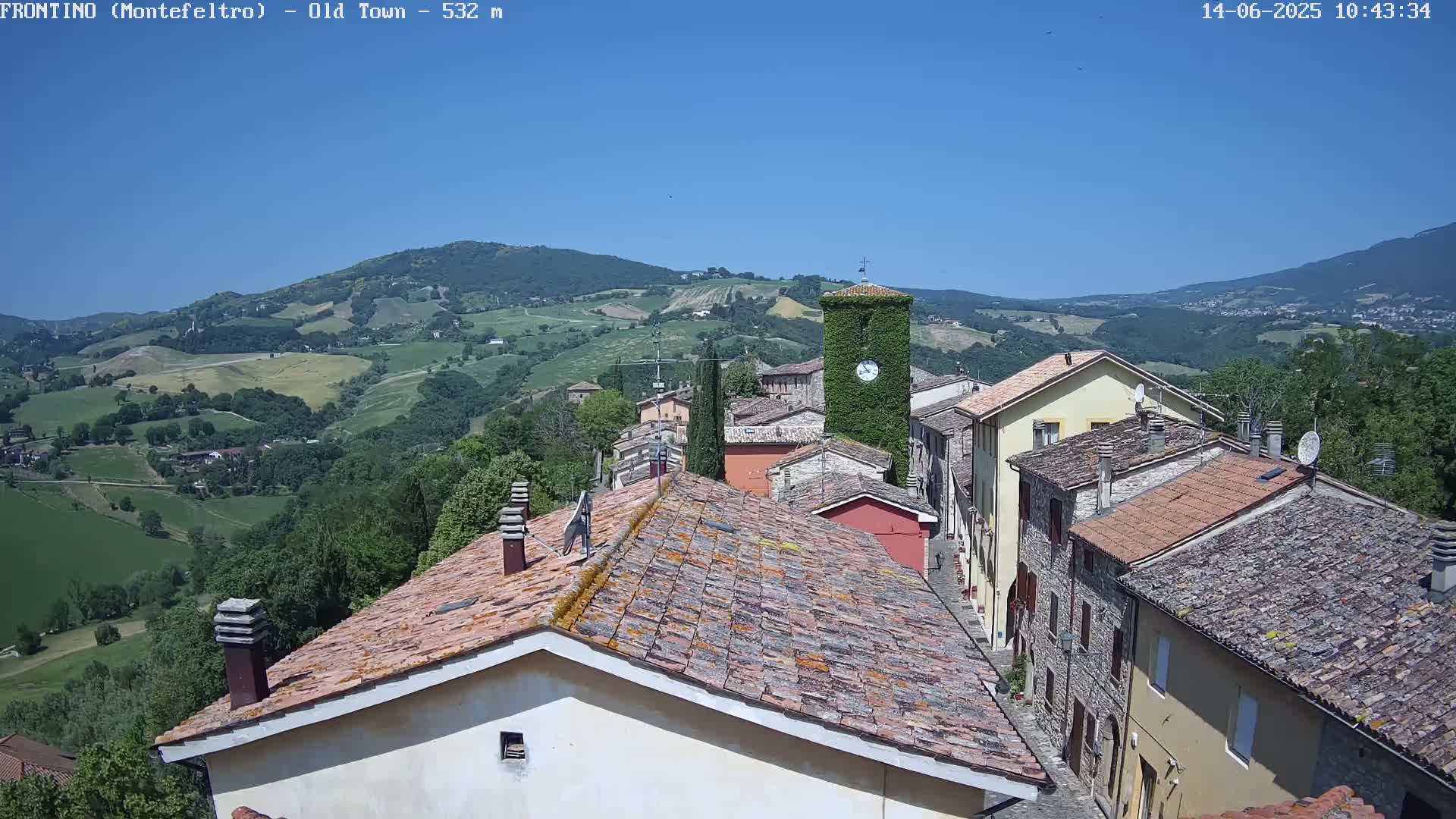 A village of stone buildings with terracotta roofs sits on a hillside overlooking a sunny valley.