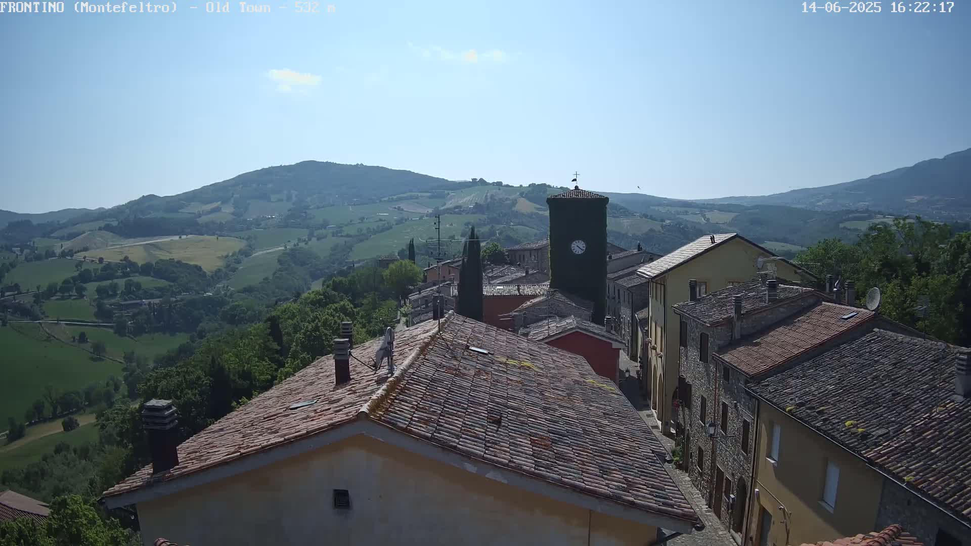 A view from above shows a small town nestled in a valley, with terracotta-tiled roofs and a clock tower, under a mostly clear blue sky.
