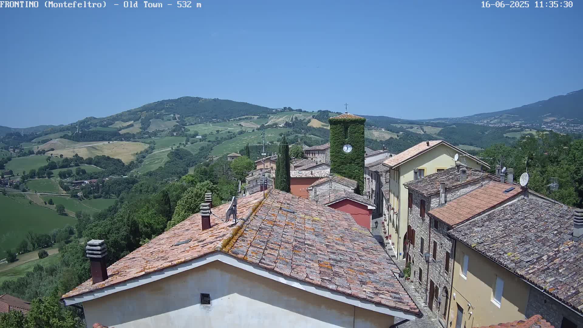 A view from atop a village of stone buildings with red-tiled roofs, overlooking a sun-drenched valley of rolling green hills under a clear blue sky.