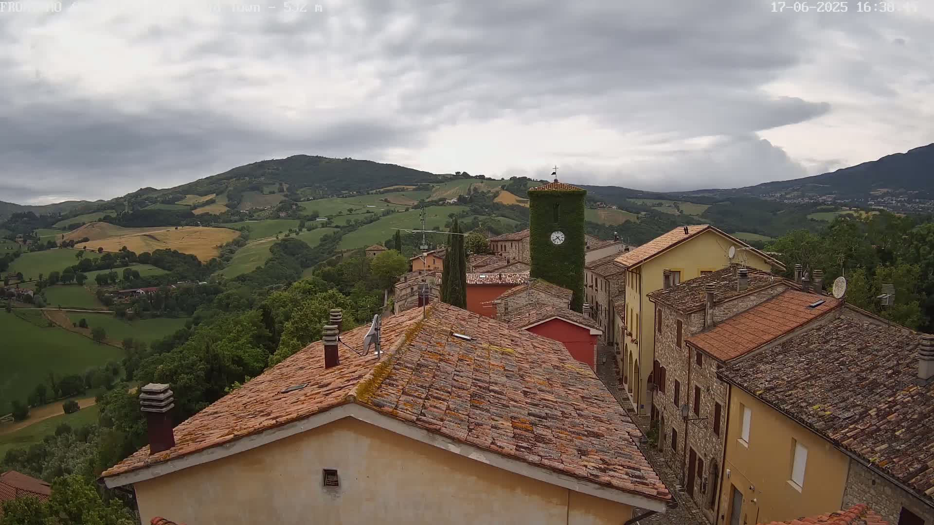 A village of stone buildings with terracotta roofs sits on a hillside overlooking a valley of rolling green fields under a cloudy sky.