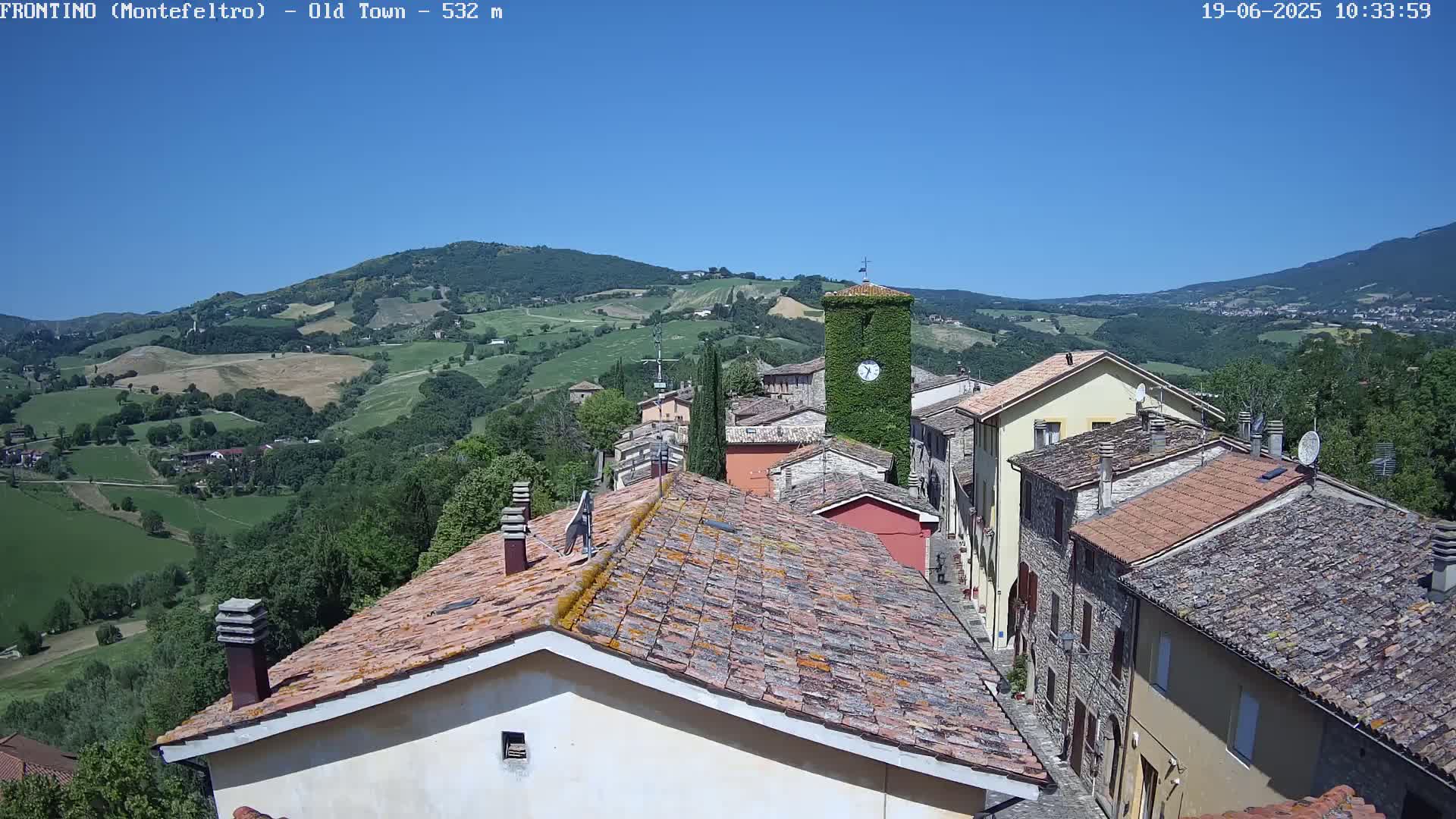 An aerial view shows a small town nestled in rolling green hills under a clear blue sky.