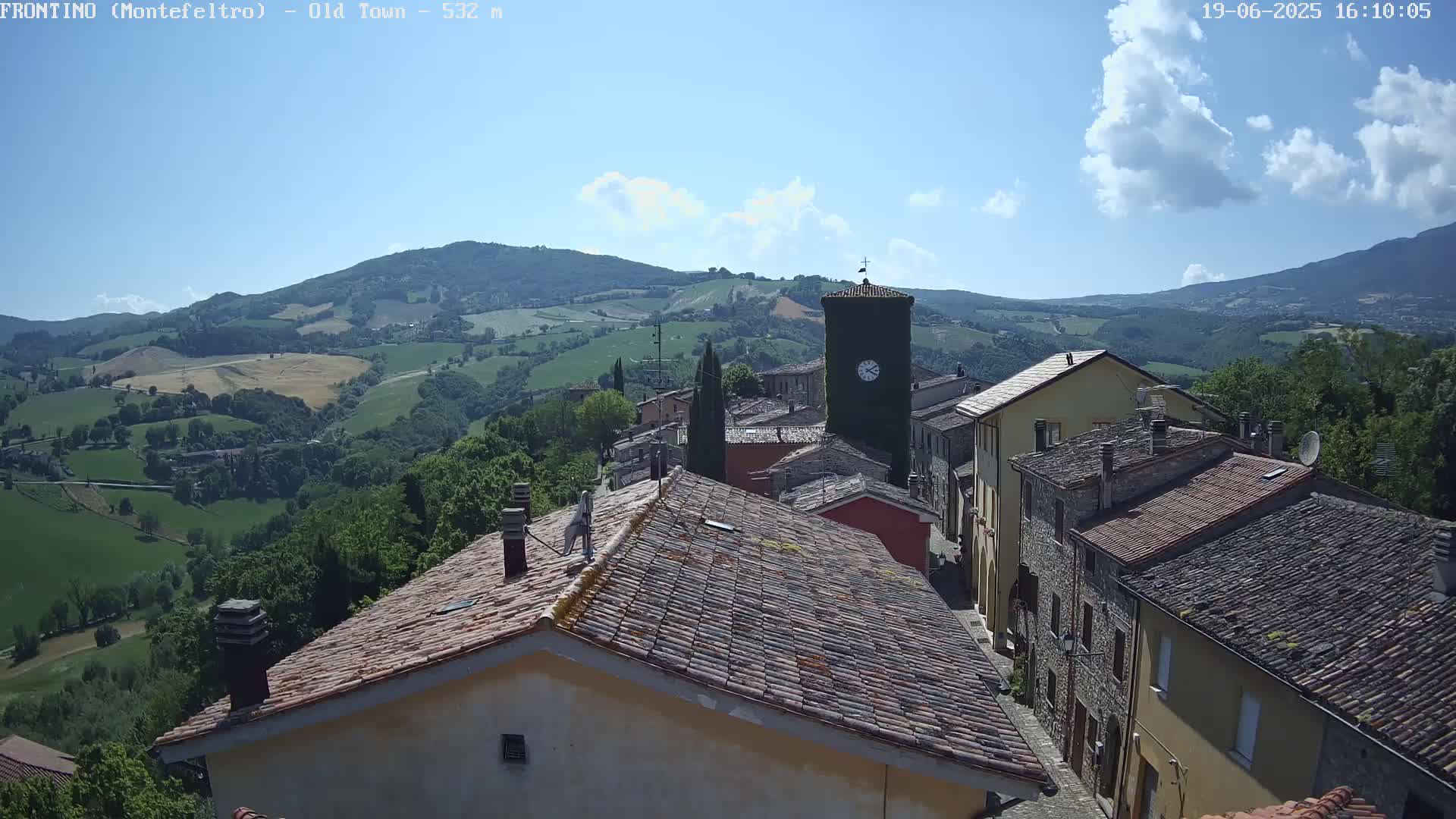 A partly cloudy day reveals a picturesque village nestled in a valley, surrounded by rolling green hills and mountains.