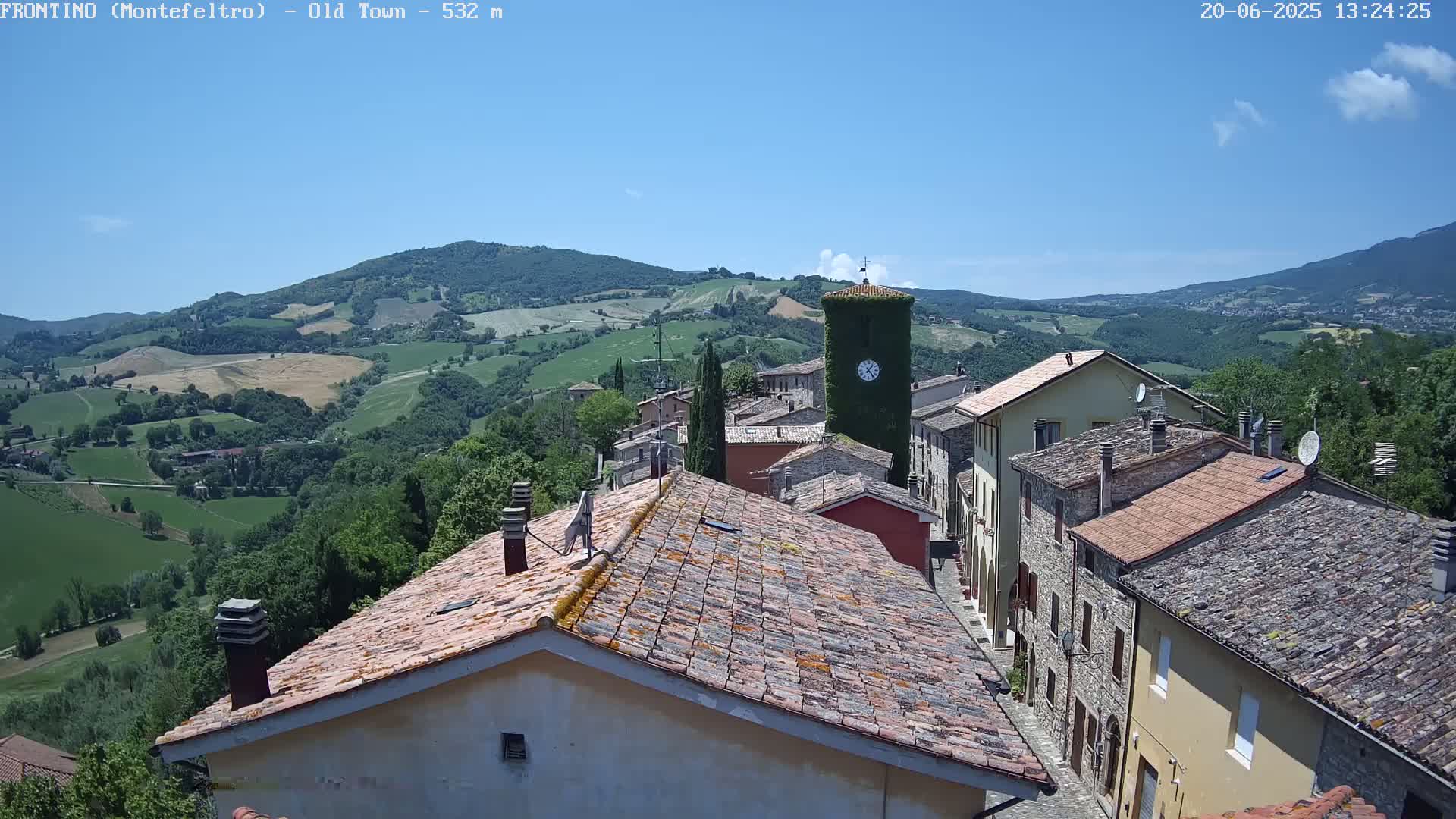 A view from above shows a small village nestled in rolling green hills under a clear blue sky.