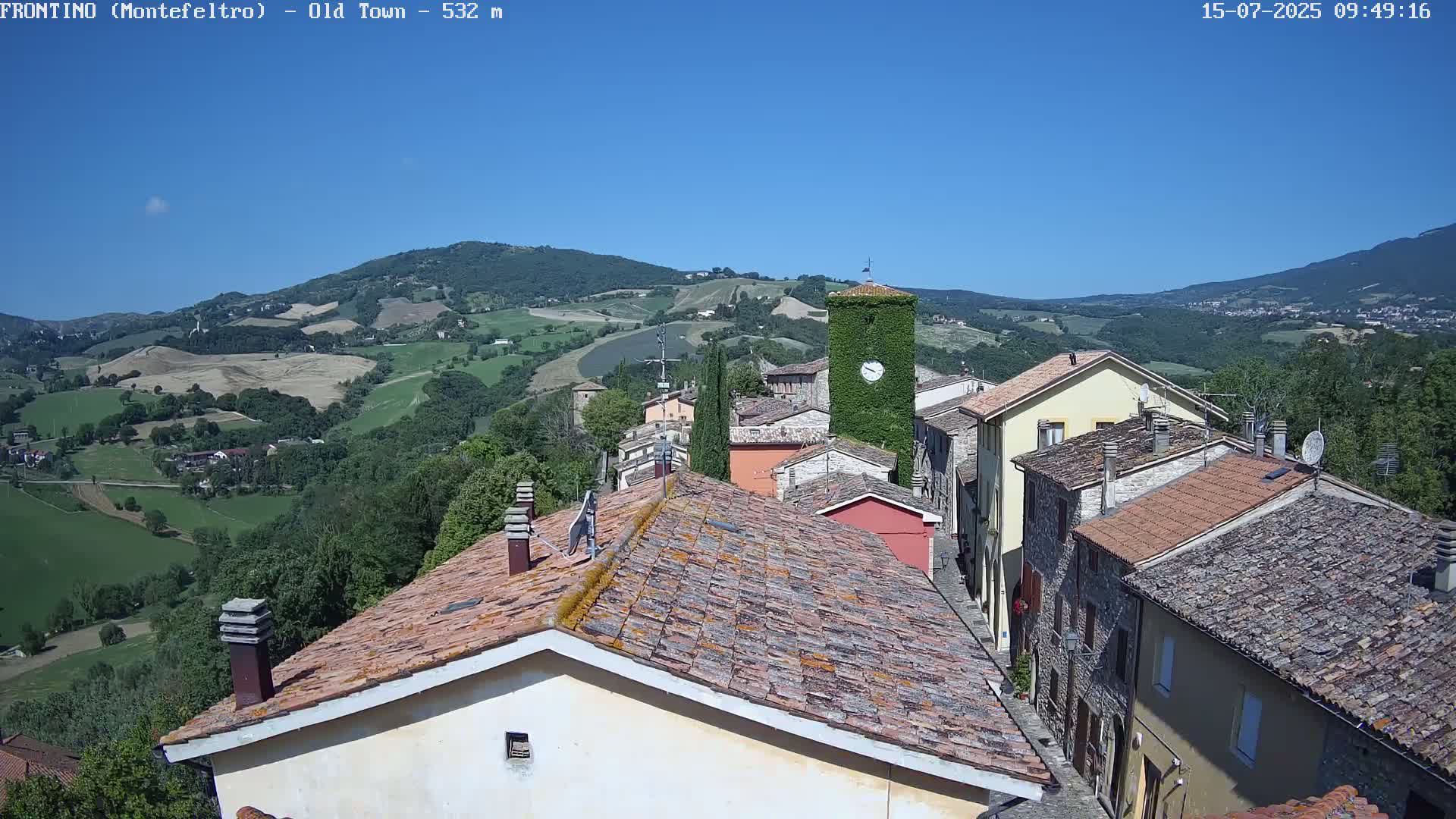A small, stone village with terracotta rooftops nestles in a verdant valley under a clear blue sky.