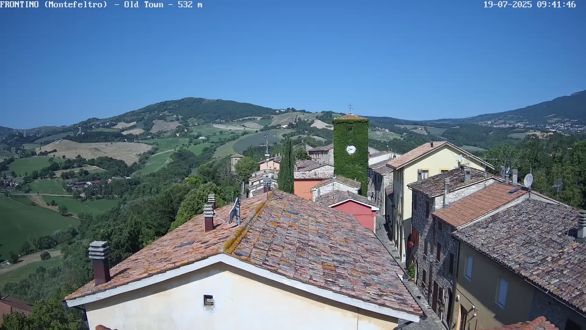 A sunlit, hilly landscape reveals a quaint town with terracotta rooftops, a clock tower covered in ivy, and a clear blue sky.