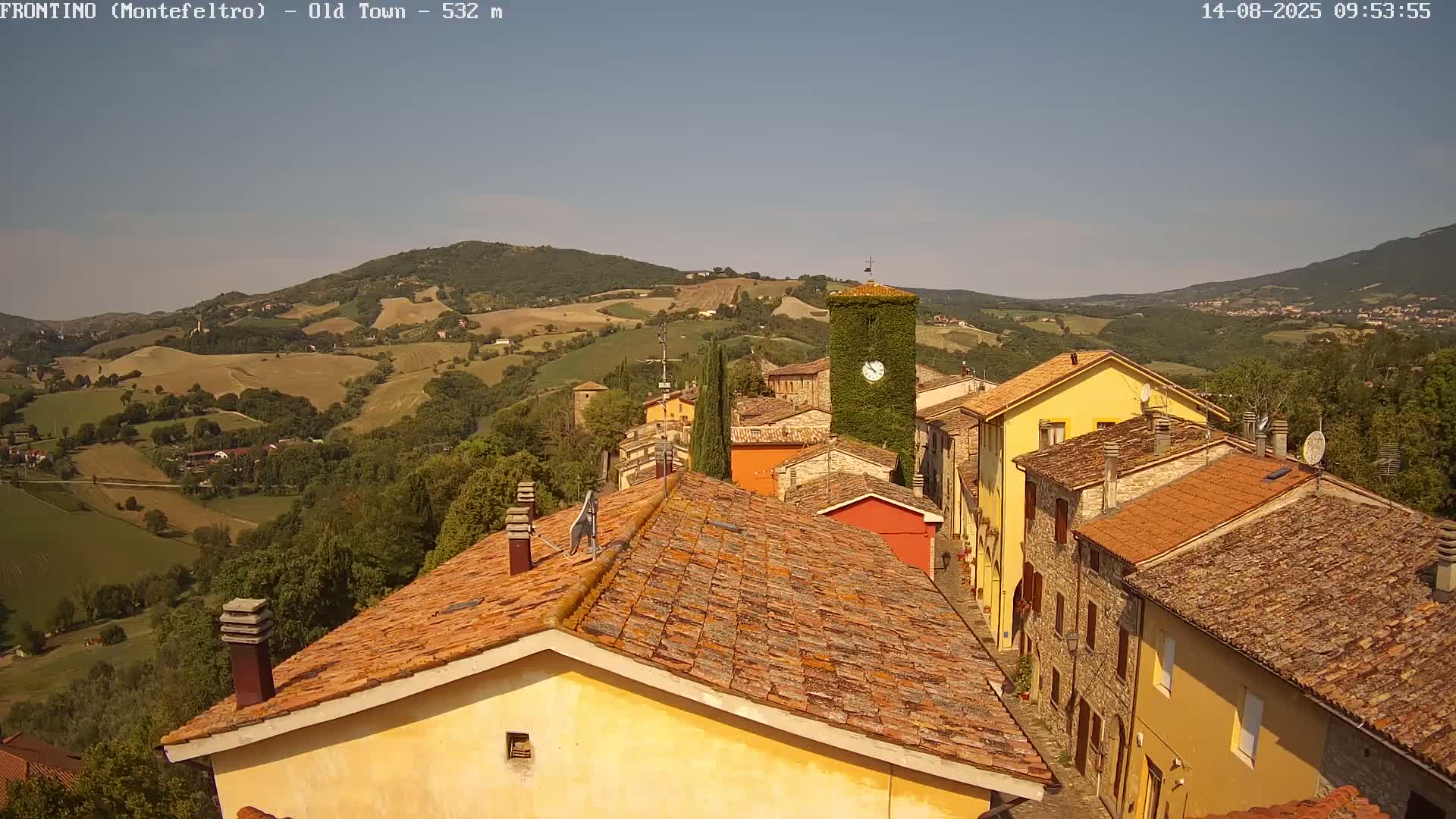 A sunlit view shows red-tiled roofs of buildings in a small town nestled in rolling green hills under a clear sky.