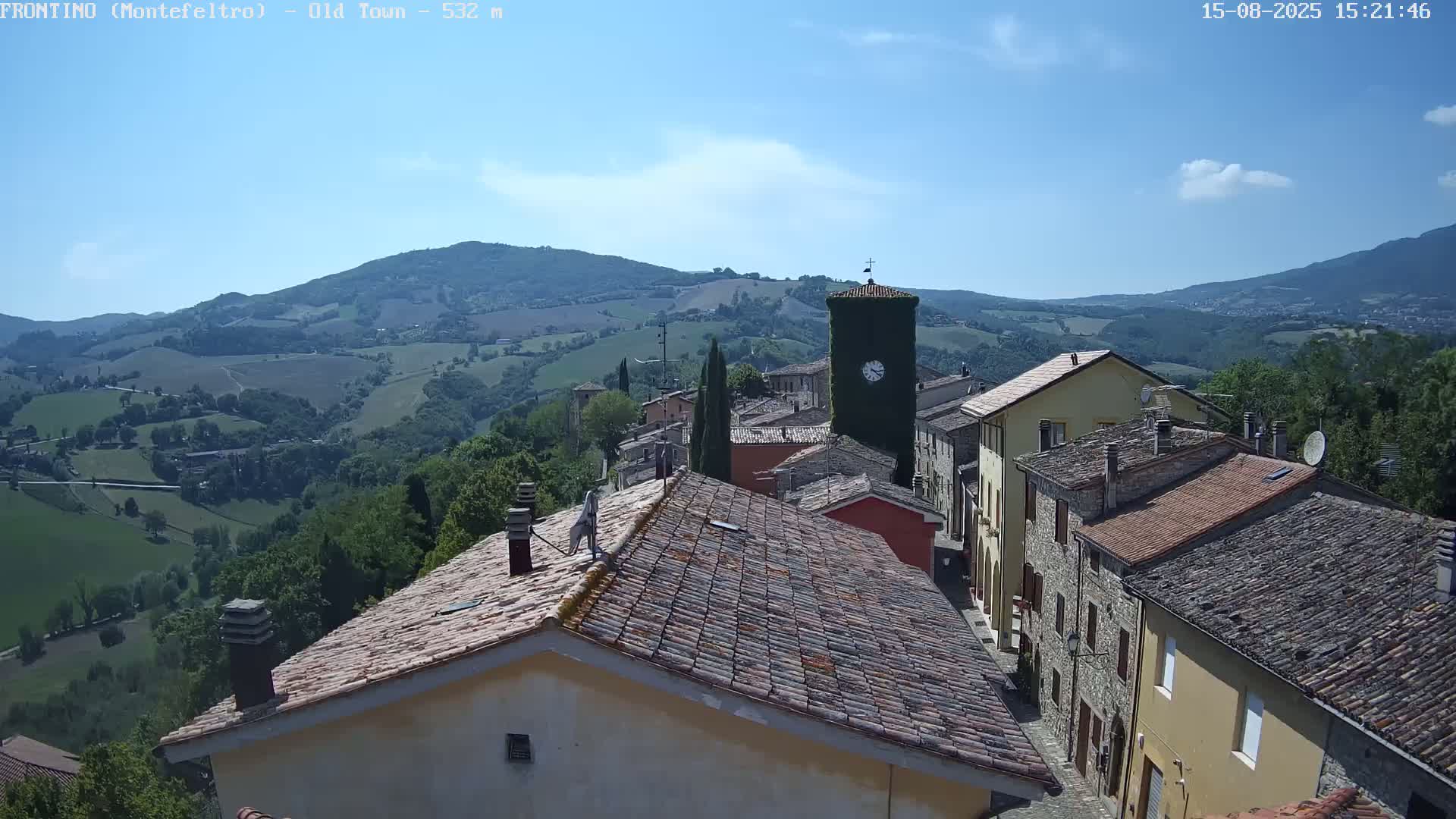 A sunlit view shows red-tiled roofs of buildings in a small town nestled in rolling green hills under a clear sky.